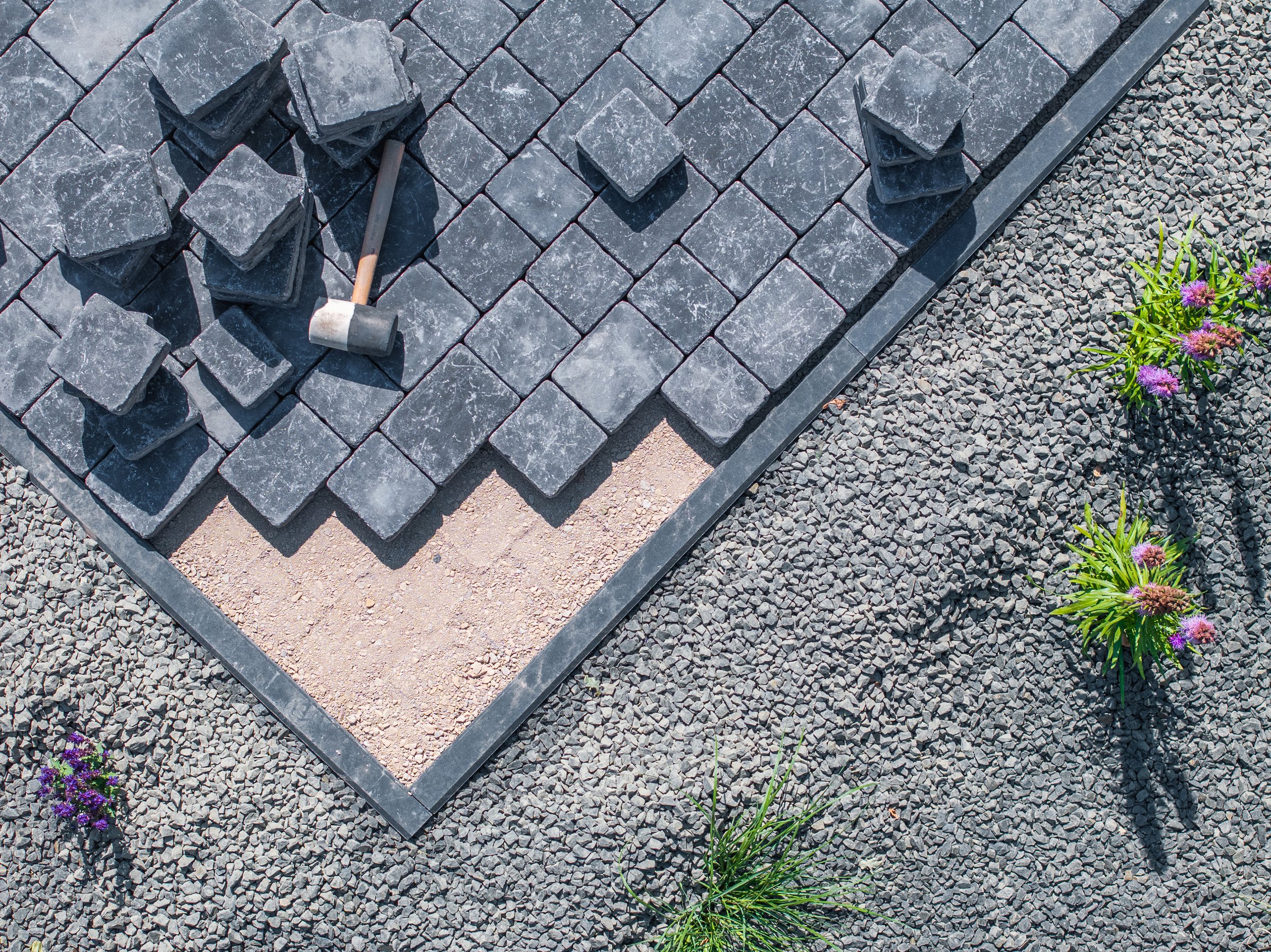 Workers are installing paving stones in a garden pathway, arranging the stones with precision under clear skies.