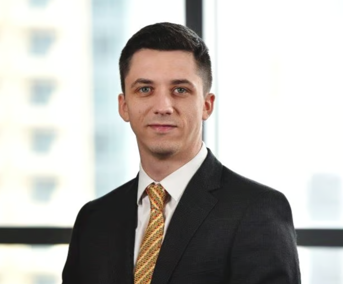 Man in a suit with a yellow tie stands in front of a blurred cityscape background.
