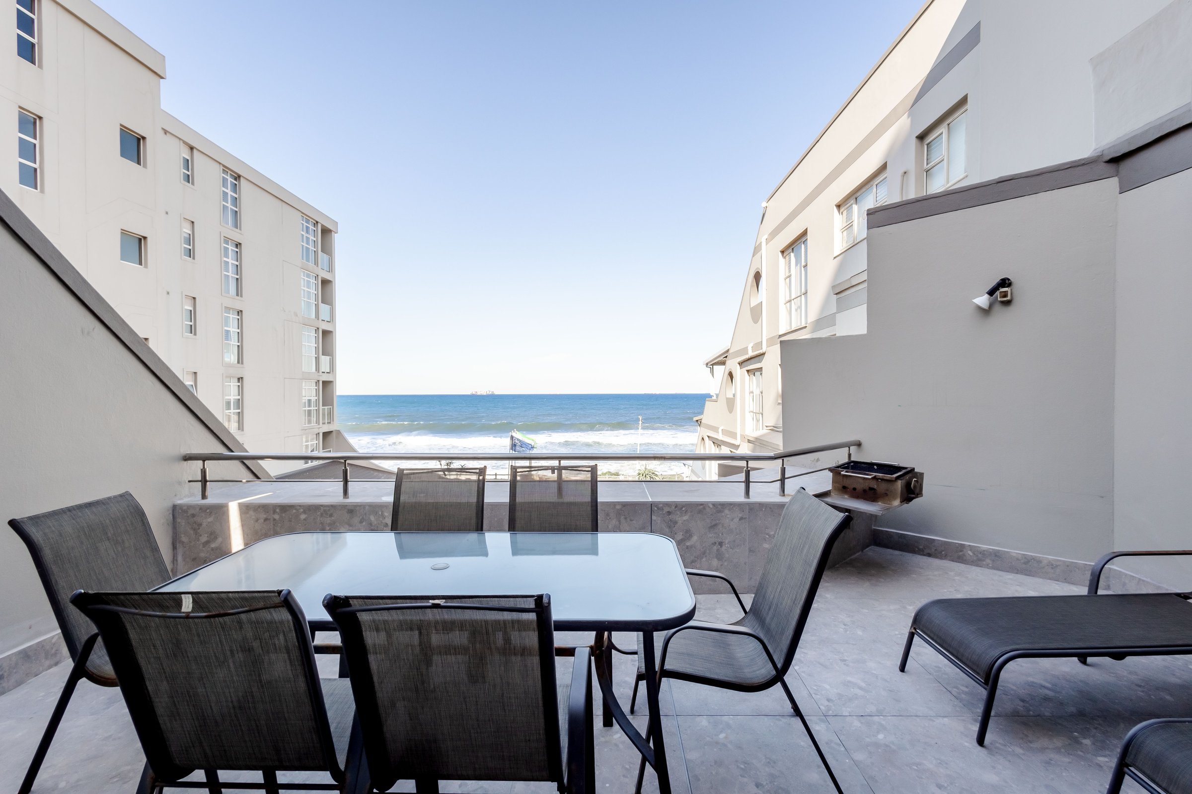 Outdoor patio with glass table, chairs, and ocean view. Surrounded by beige buildings, clear sky in the background.