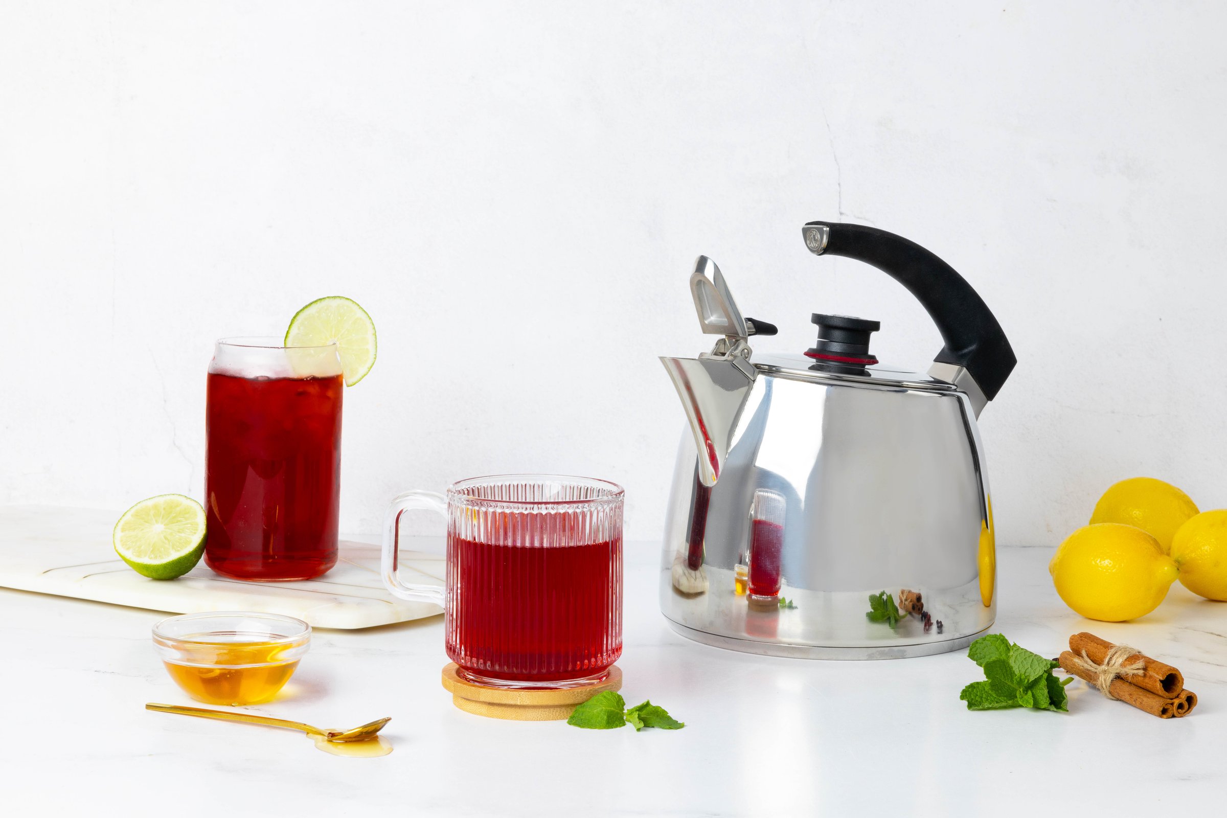 Tea setup with a stainless steel kettle, two glasses of red tea with lime, lemons, cinnamon, honey, and mint leaves.
