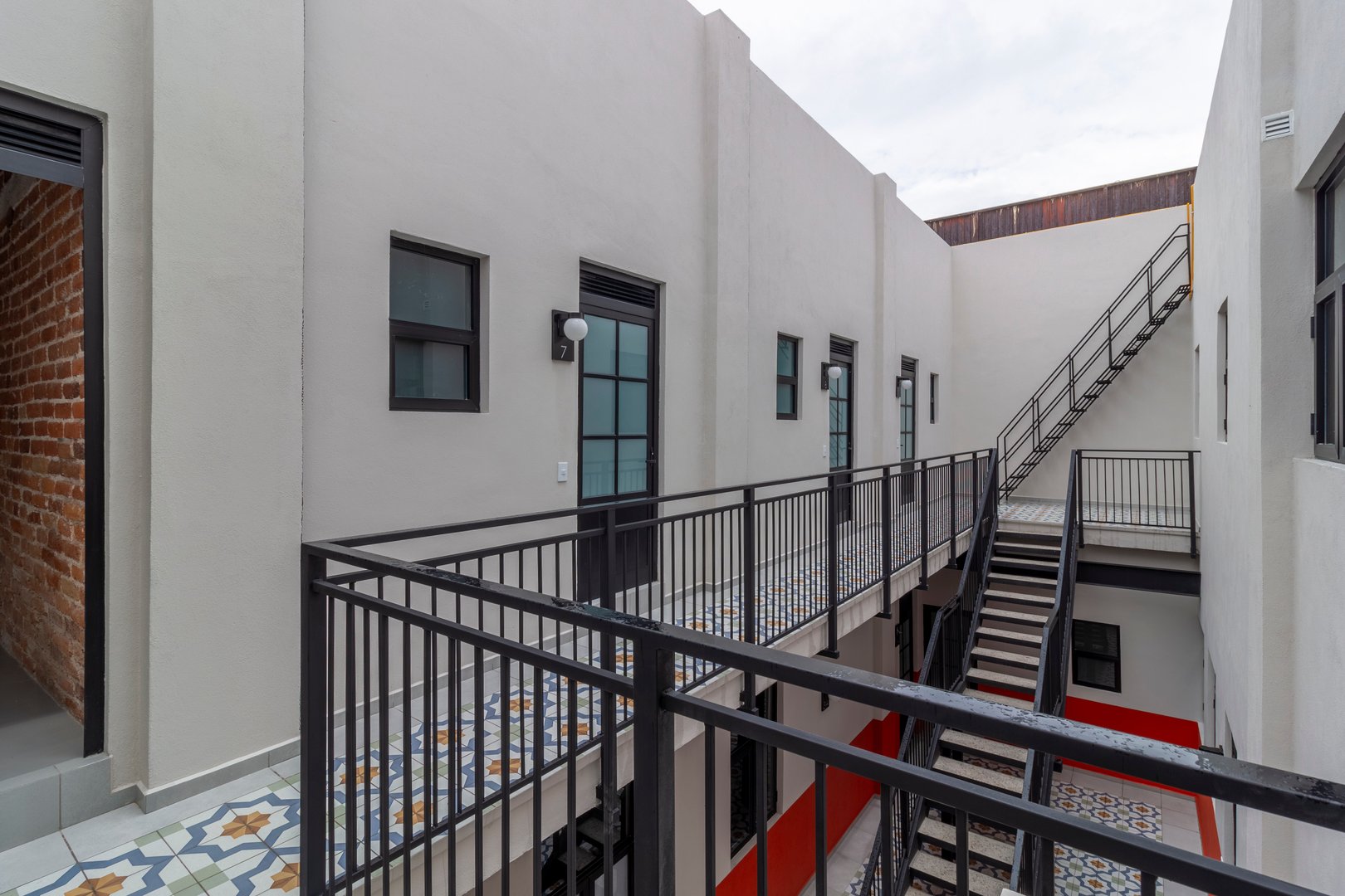 Modern apartment building with black railings, stairs, and patterned tiles on the walkway, featuring light gray walls.
