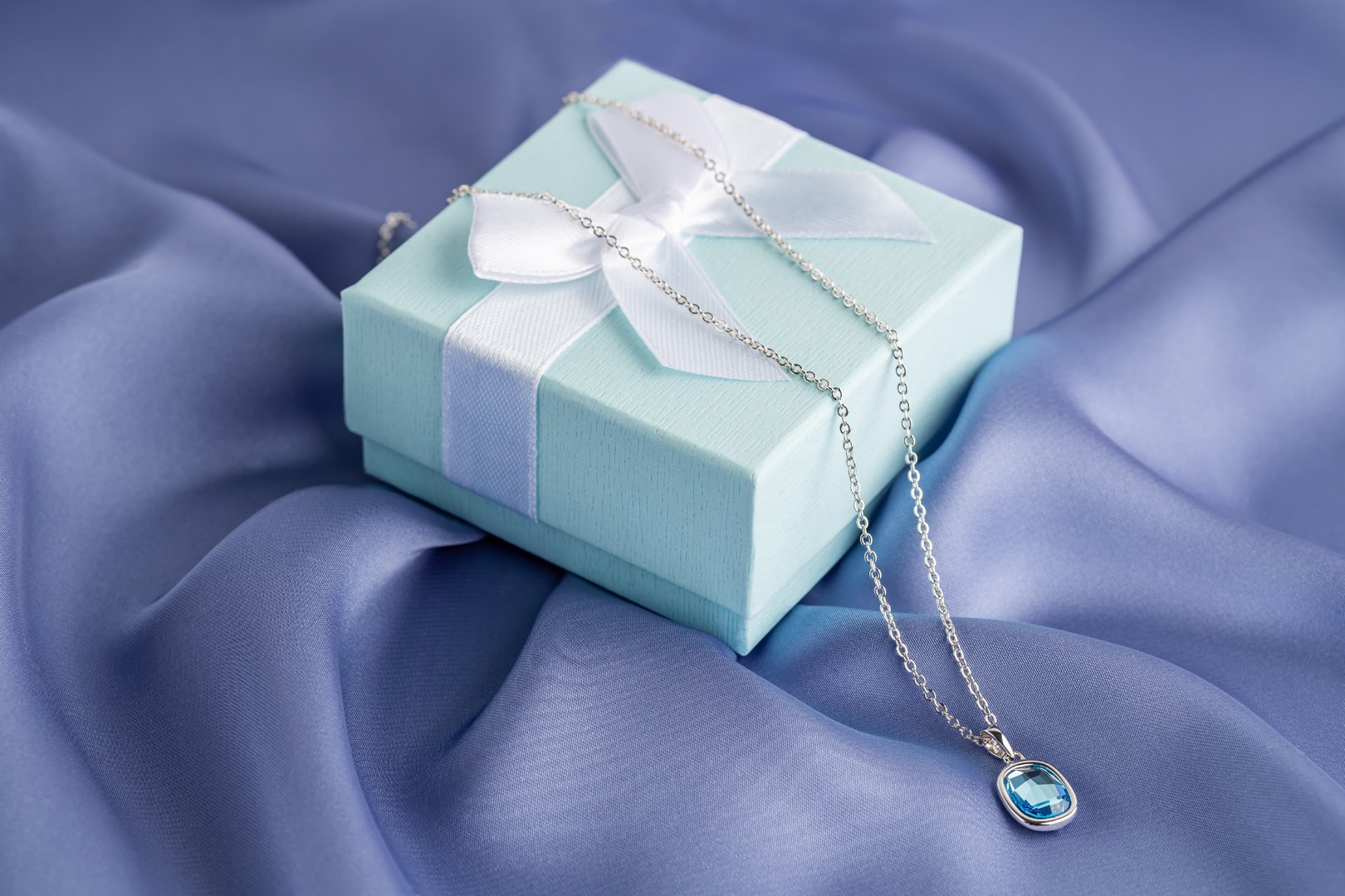 Green gift box with white ribbon and necklace with medallion with blue gemstone on the blue silk background.