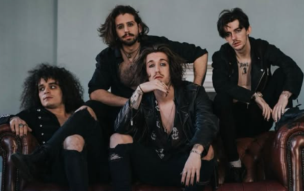 Four men in rock attire sitting and posing on a leather couch, with long hair and leather jackets, exuding a rock band vibe.