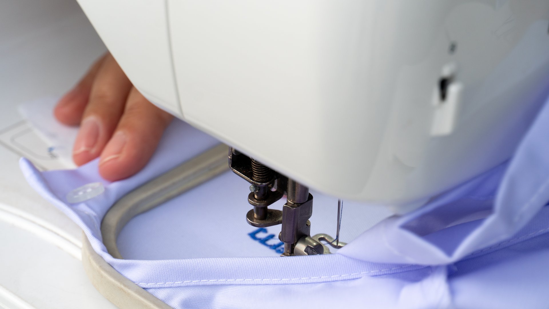 Person hand guiding light blue fabric under the needle of a white embroidery machine. The machine is actively stitching a blue design onto the fabric held taut in a hoop.