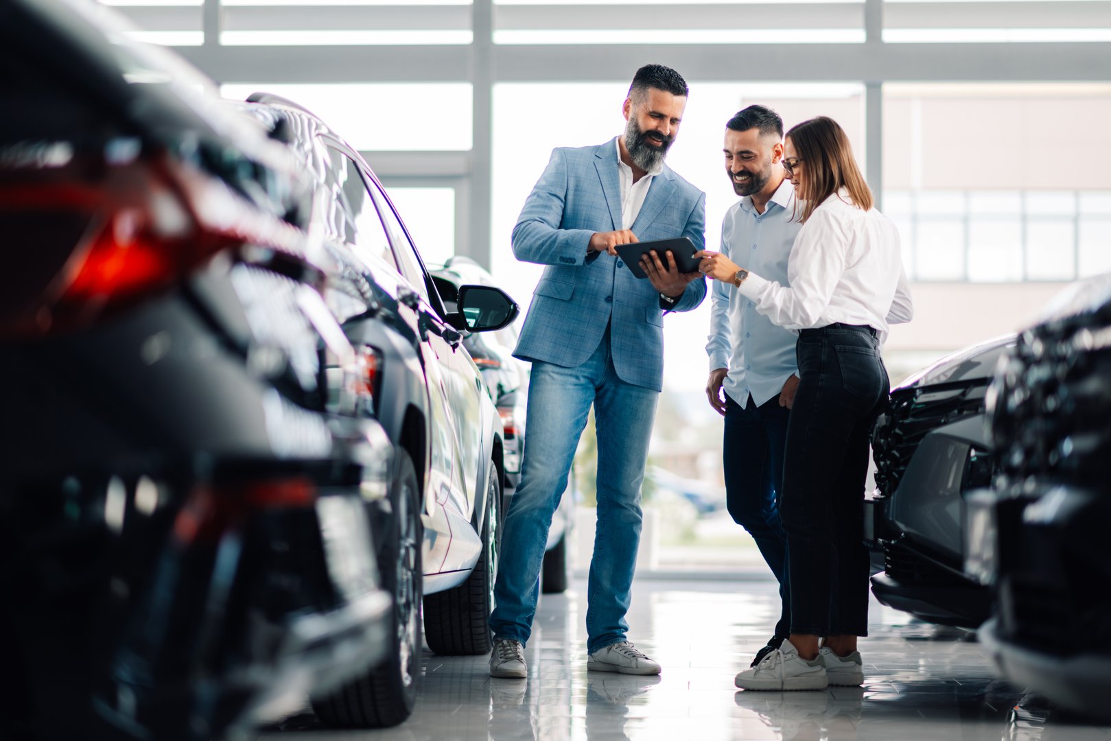 A car dealership scene with three people discussing vehicle choices while looking at a tablet, surrounded by various cars in a bright showroom setting.