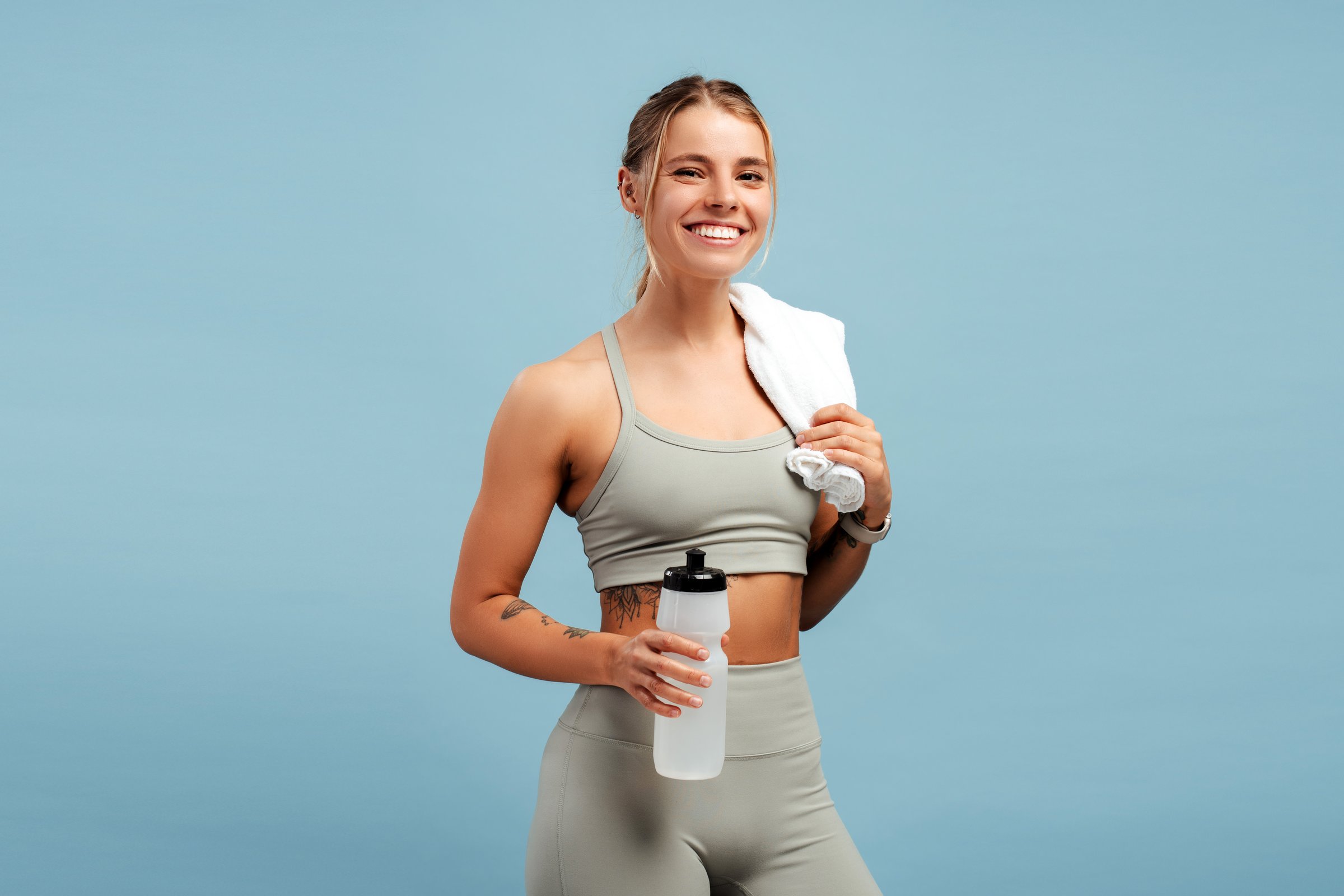 Happy fit pretty woman holding bottle of water and white towel, looking at camera, isolated on blue background. Sports, diet, fitness concept