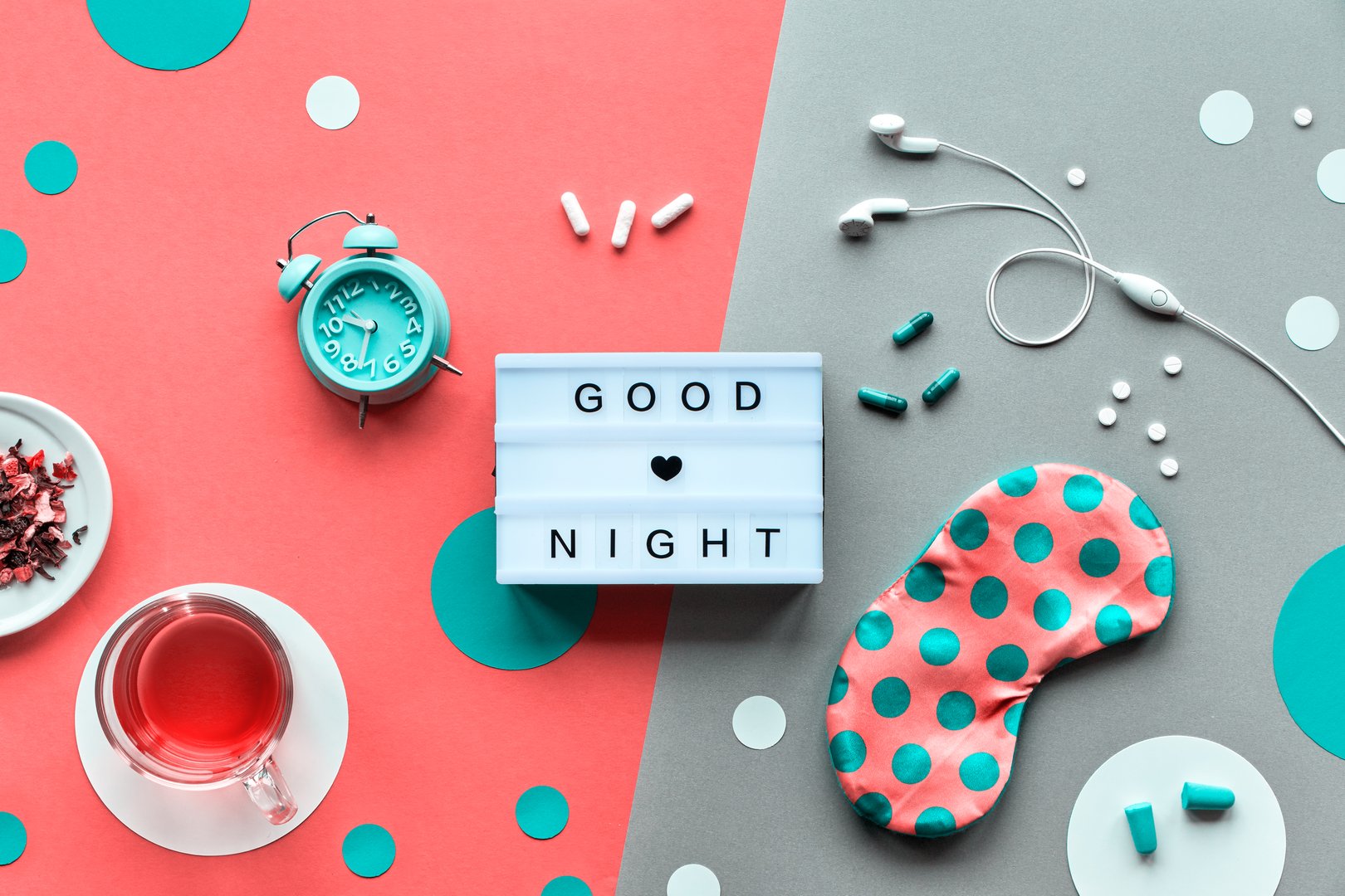 Text 'Good night' on trendy lightbox. Pink sleeping mask with polka dots, alarm clock, earphones and earplugs. Pills, capsules and calming tea. Flat lay, two tone orange coral and silver background.