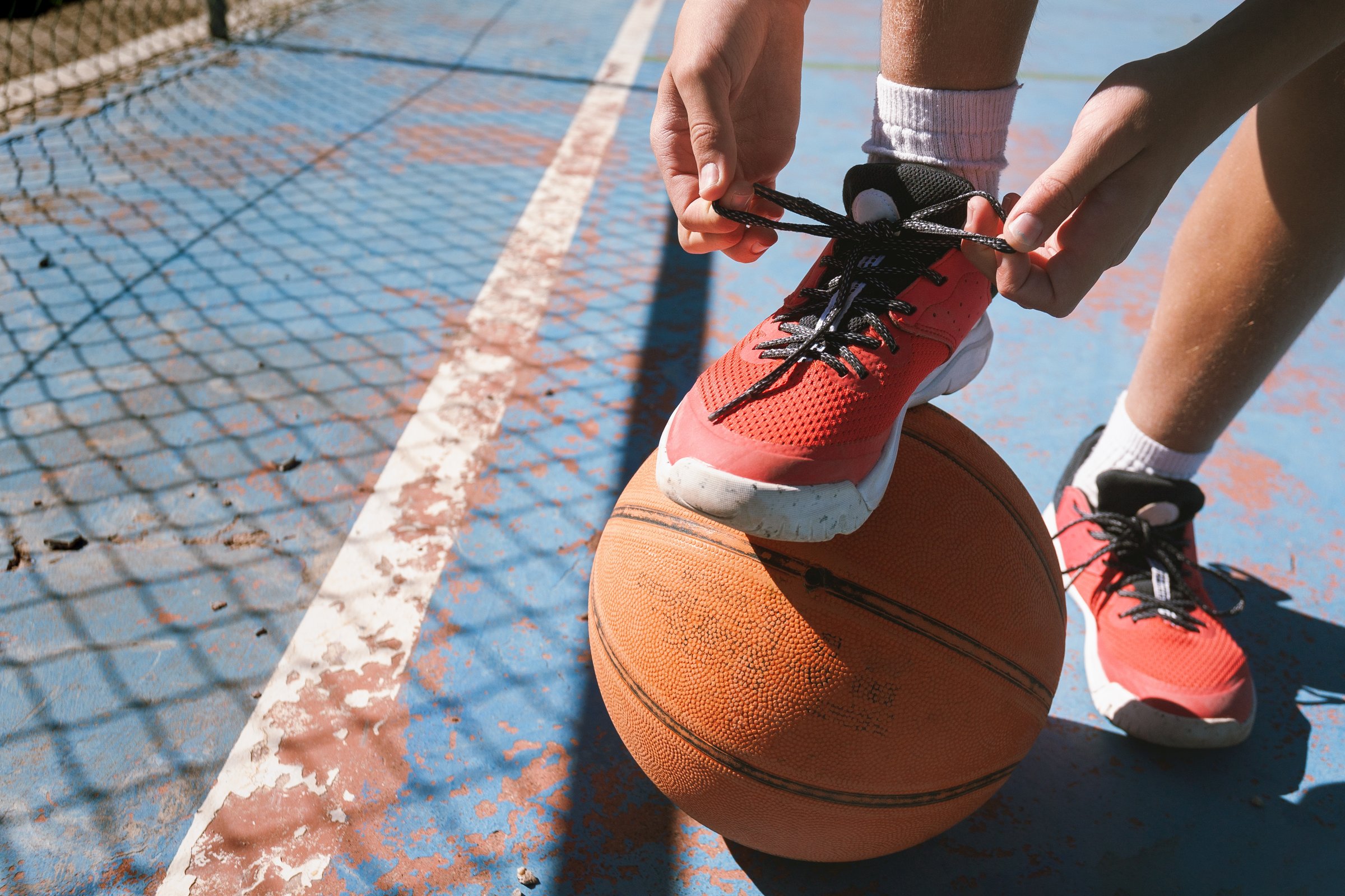 A person is tying their shoe while standing next to a basketball. The scene is set on a court, with a basketball resting on the ground. The person appears to be focused on their shoe