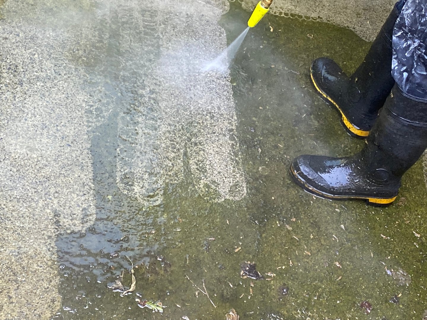 A person wearing rubber boots cleans the cement surface using power washing machine