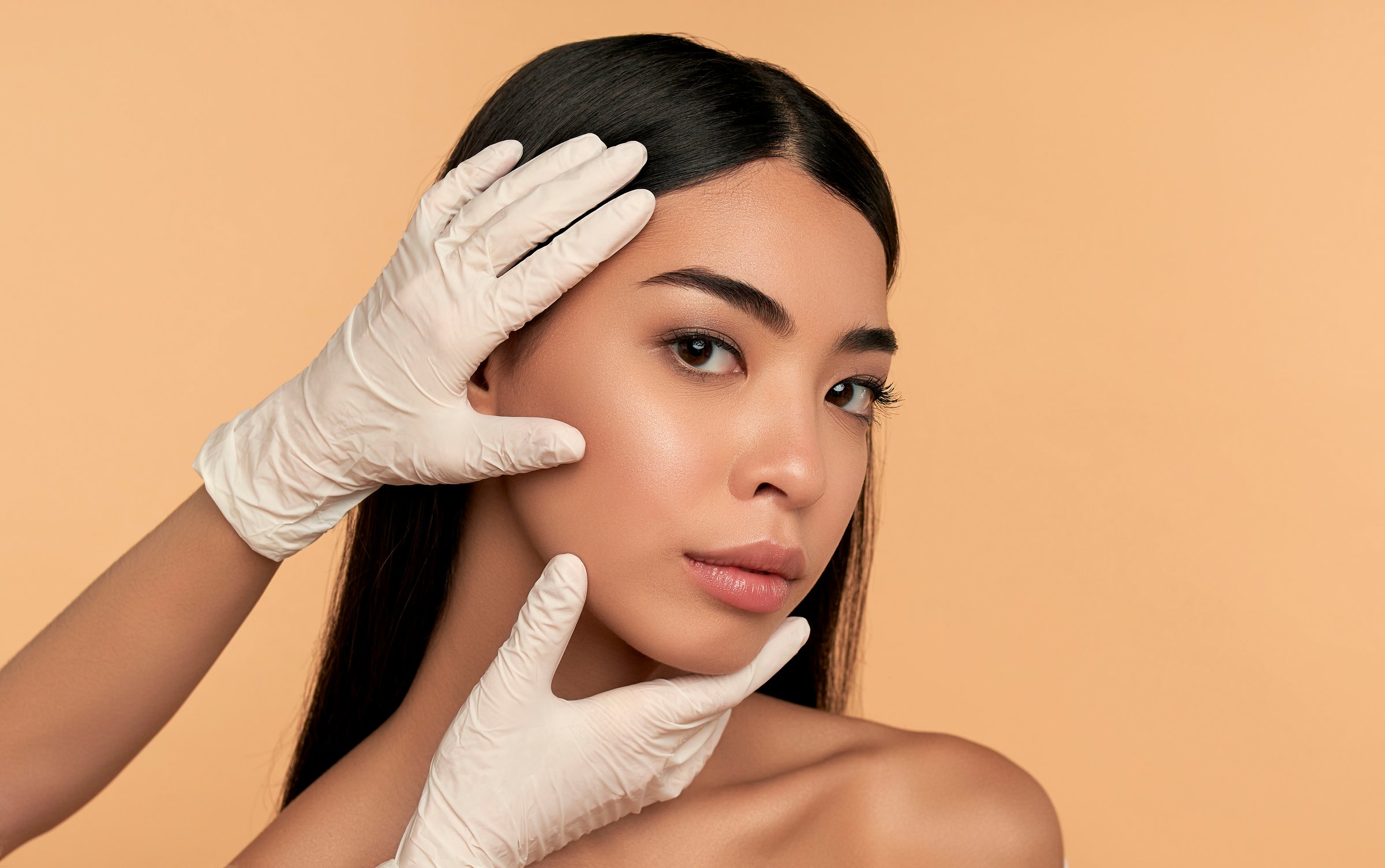 Young Asian woman with clean radiant skin gets botox injections for contour tightening, lip augmentation on a beige background. Spa care, facial skin care, beauty cosmetology.