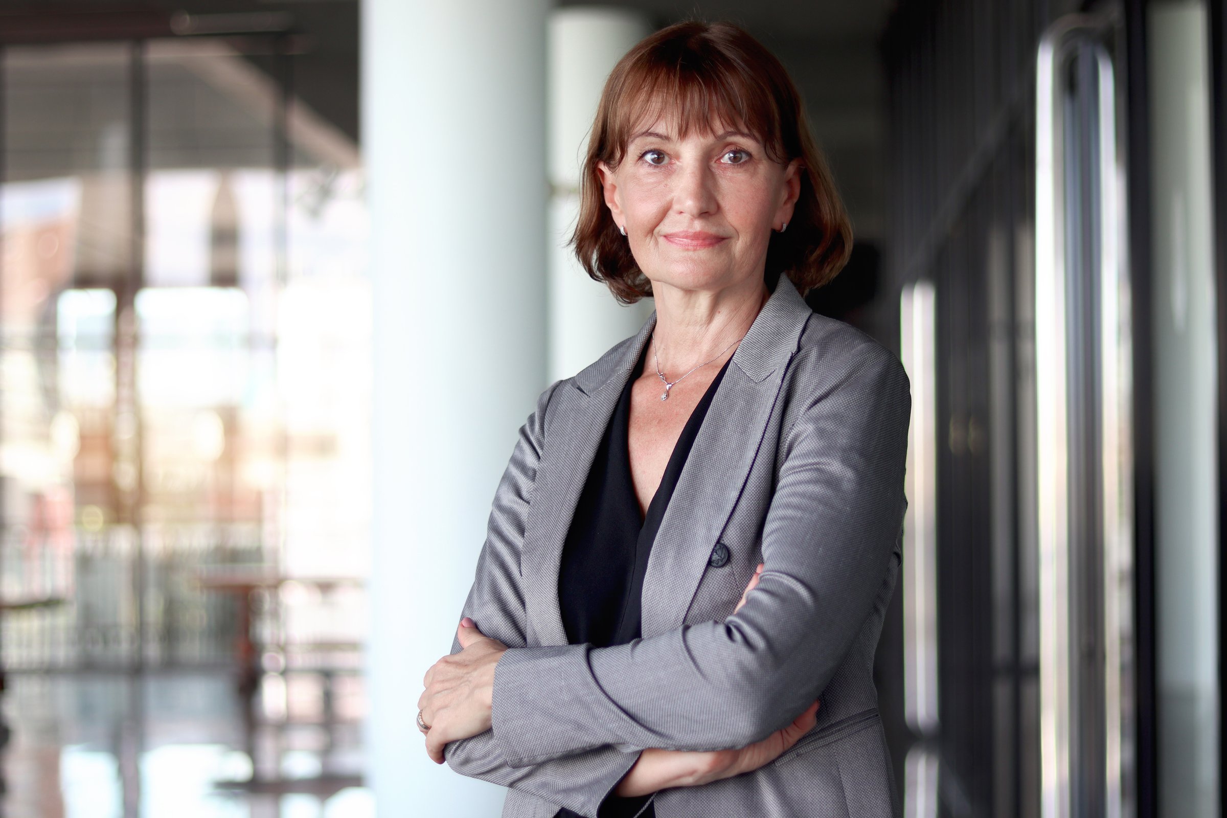 Happy smiling senior businesswoman in suit standing with arms crossed in front of office. Elderly successful female boss working at workplace. Portrait of Caucasian white mature woman worker.