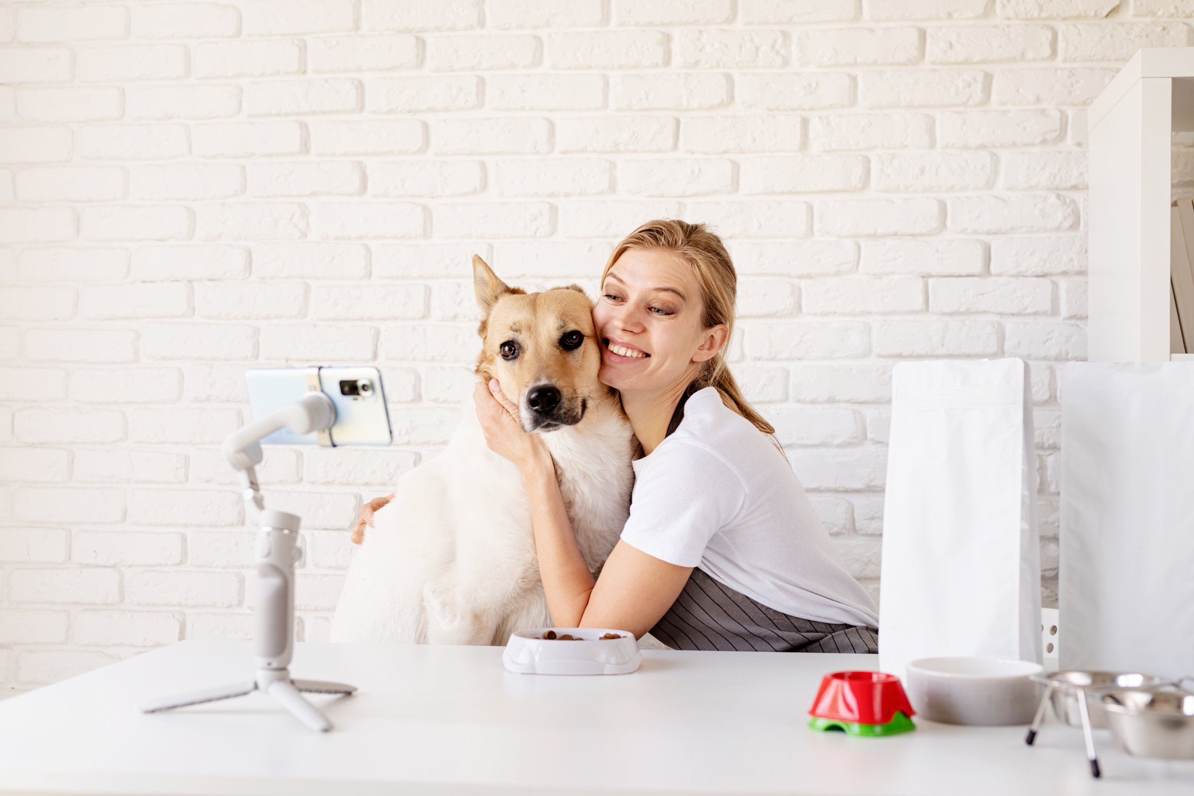 Smiling young woman blogger influencer at home with her mixed breed dog. Woman speaking about dog food looking at camera talking, making video chat, conference call lifestyle blog vlog.