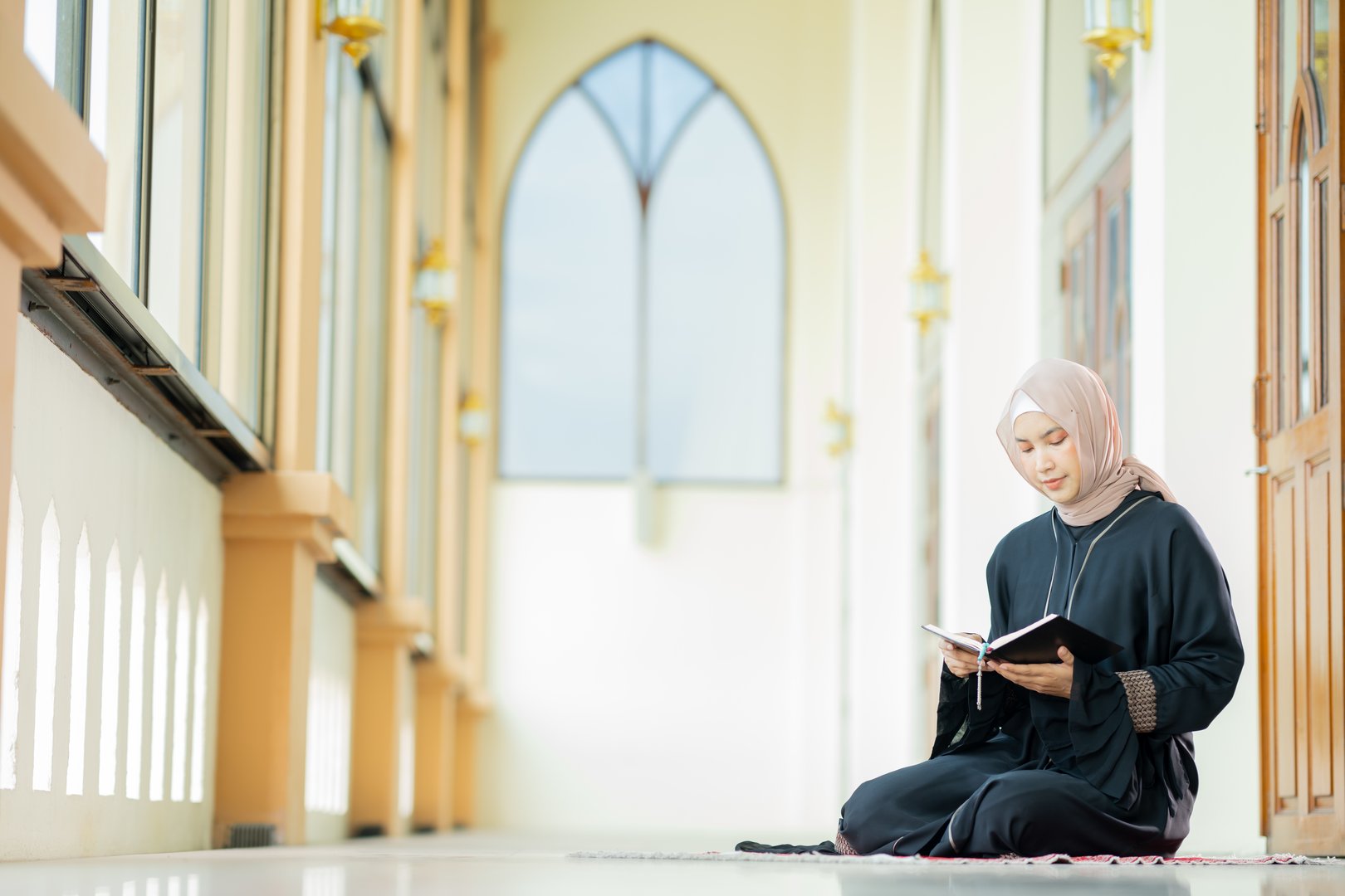 The image of an Asian Muslim woman in the Islamic religion in hijab in cream color. reading the Quran and having a happy Staying in a beautiful mosque, Arabic word translation: The Holy Al Quran.