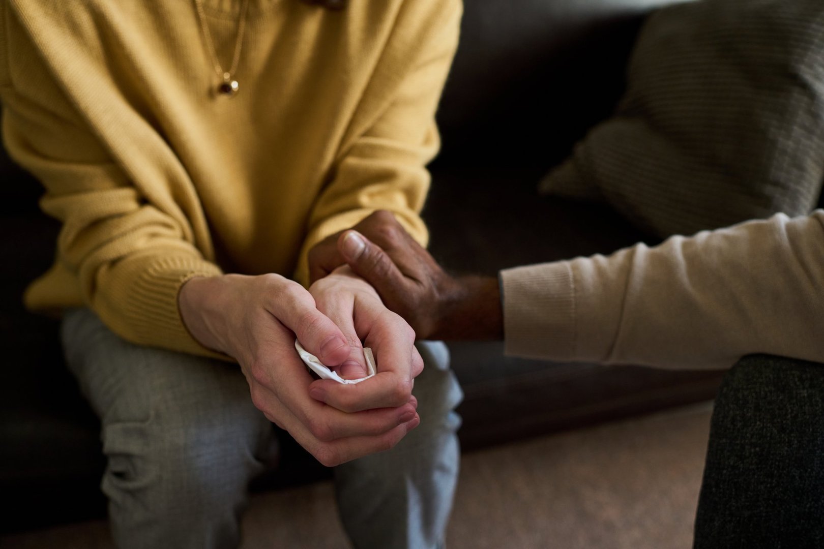 Caucasian young adult woman sitting with hands clasped holding tissue, Black middle aged man gently touching her arm offering comfort, both seated on sofa in indoor setting