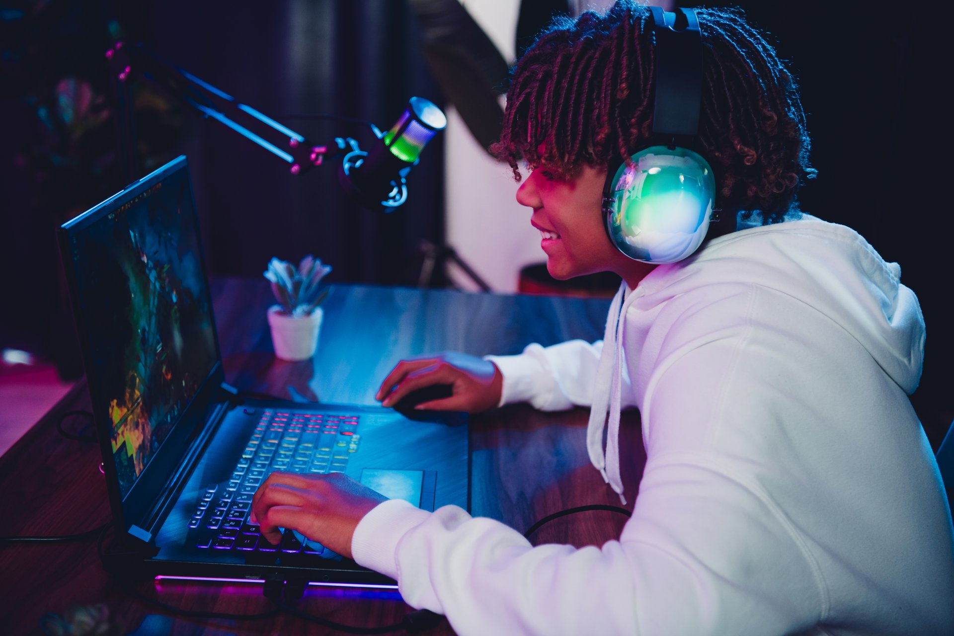 Male teen enjoying an evening gaming and streaming experience on his laptop with glowing headphones and a microphone in a vibrant neon-lit environment