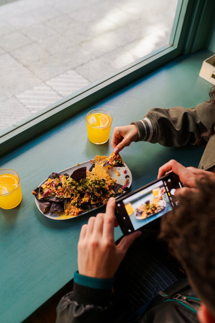 Food blogger photographing nachos with guacamole and orange juice at a restaurant table by the window