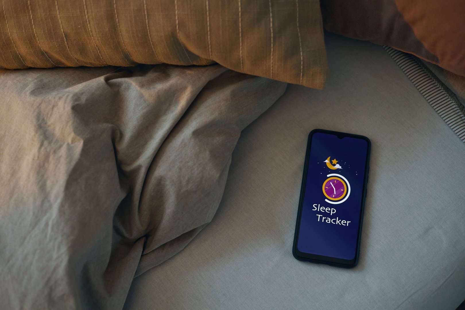 Smartphone showing sleep tracking app, resting on bed in warm lighting. Pillow and blanket creating a cozy atmosphere