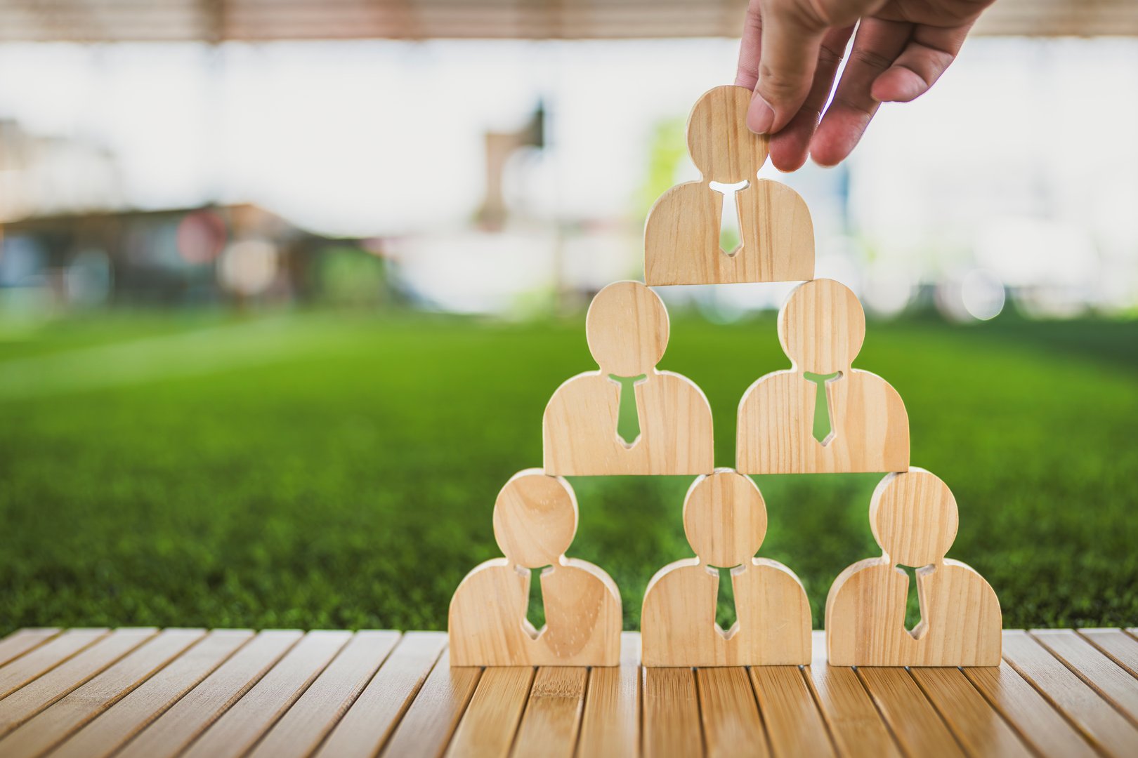 Wooden human icons forming a pyramid structure with a hand placing the top piece, symbolizing leadership, team building, business hierarchy, management strategy, and organizational structure.