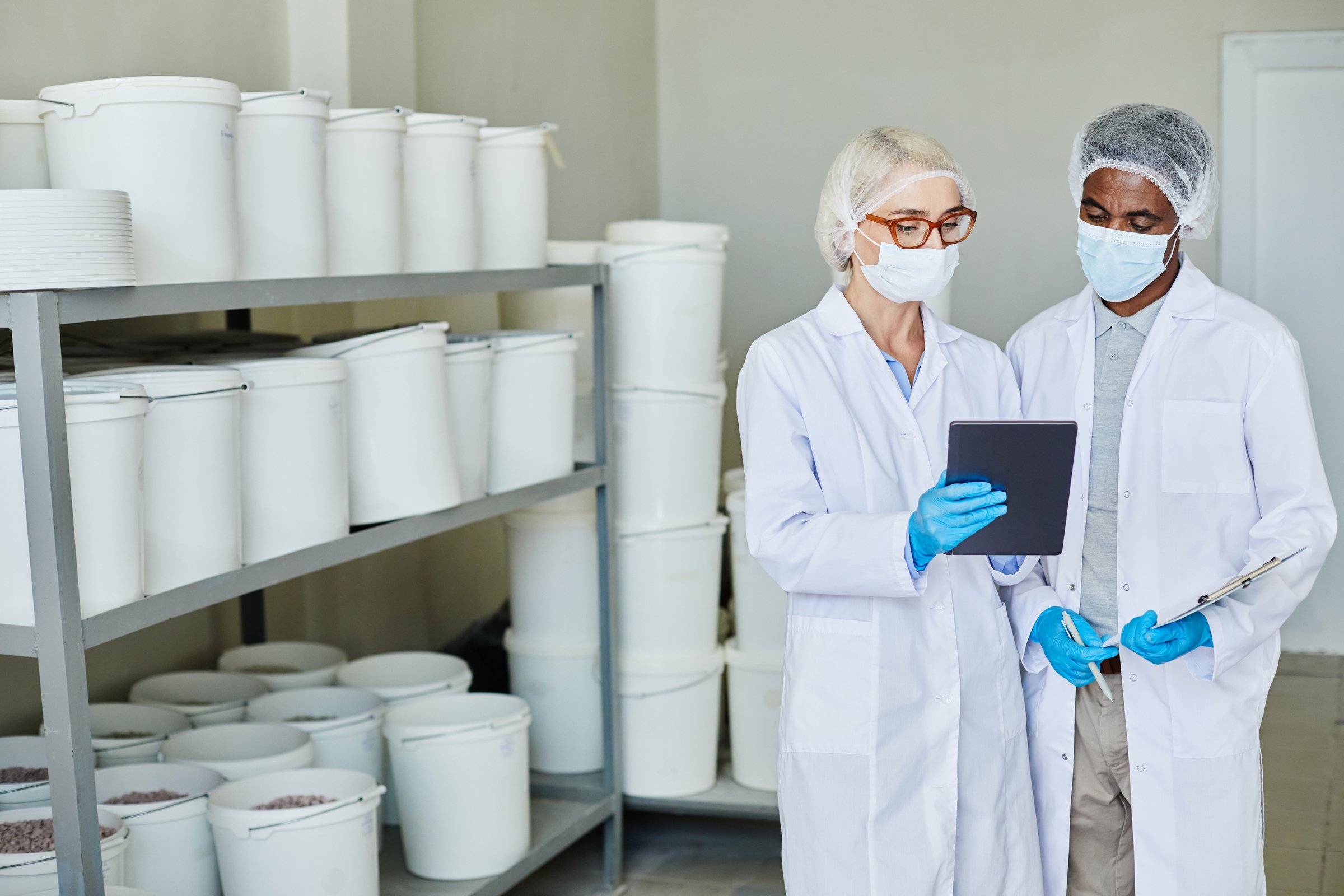 Medium shot of two supervisors in protective masks and lab coats using digital tablet to take inventory of pharmaceutical products stored in large plastic containers at manufacturing plant, copy space