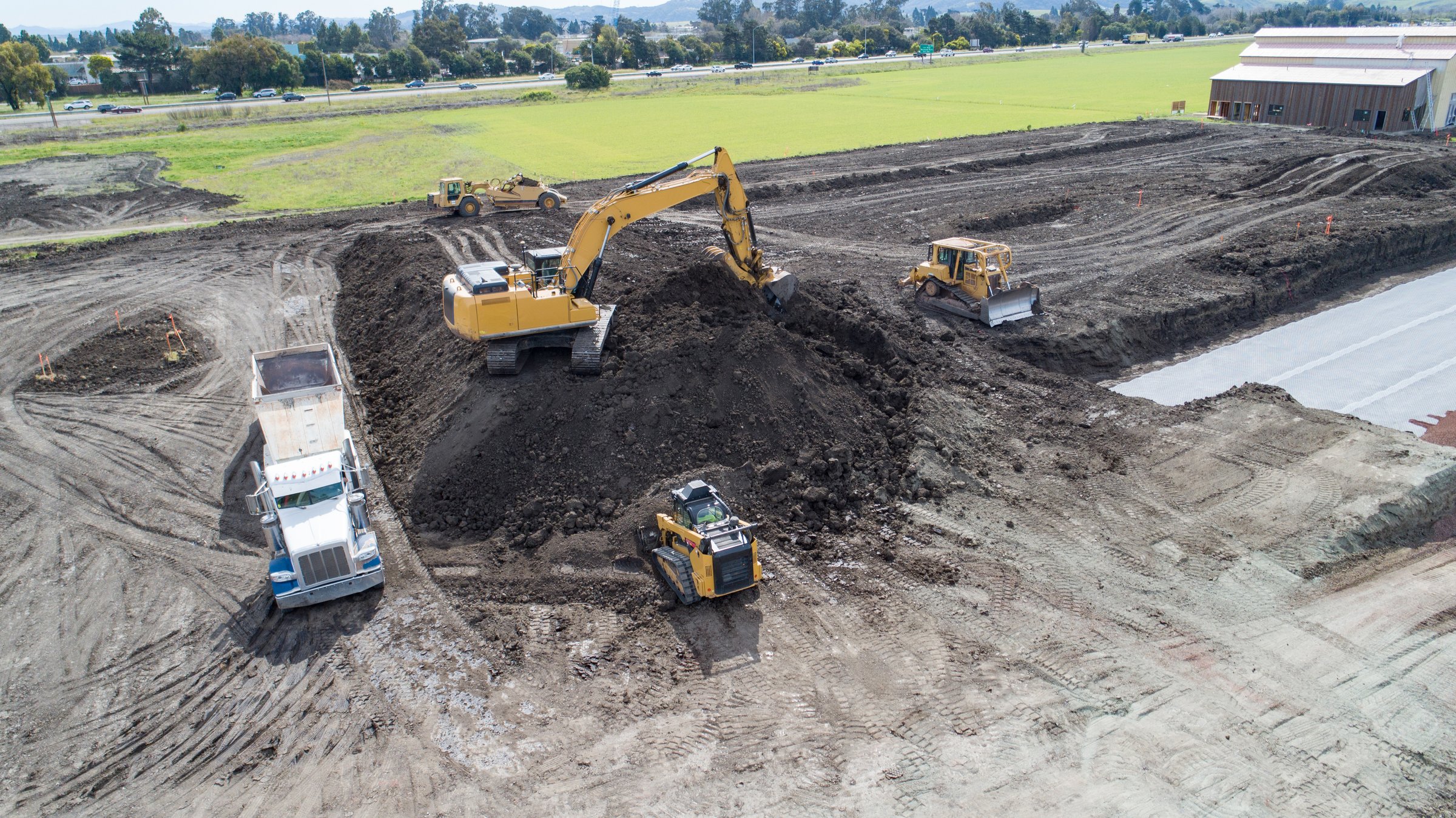 Yellow and black heavy equipment loading dirt into dump truck on construction site. Excavator, Skip Loader, Bulldozer, and dump truck are moving large pile of dark dirt