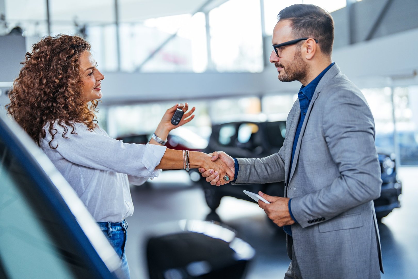 A smiling customer receives car keys from a professional salesman in a bright showroom. The scene conveys satisfaction and successful business dealings in an automotive retail environment.