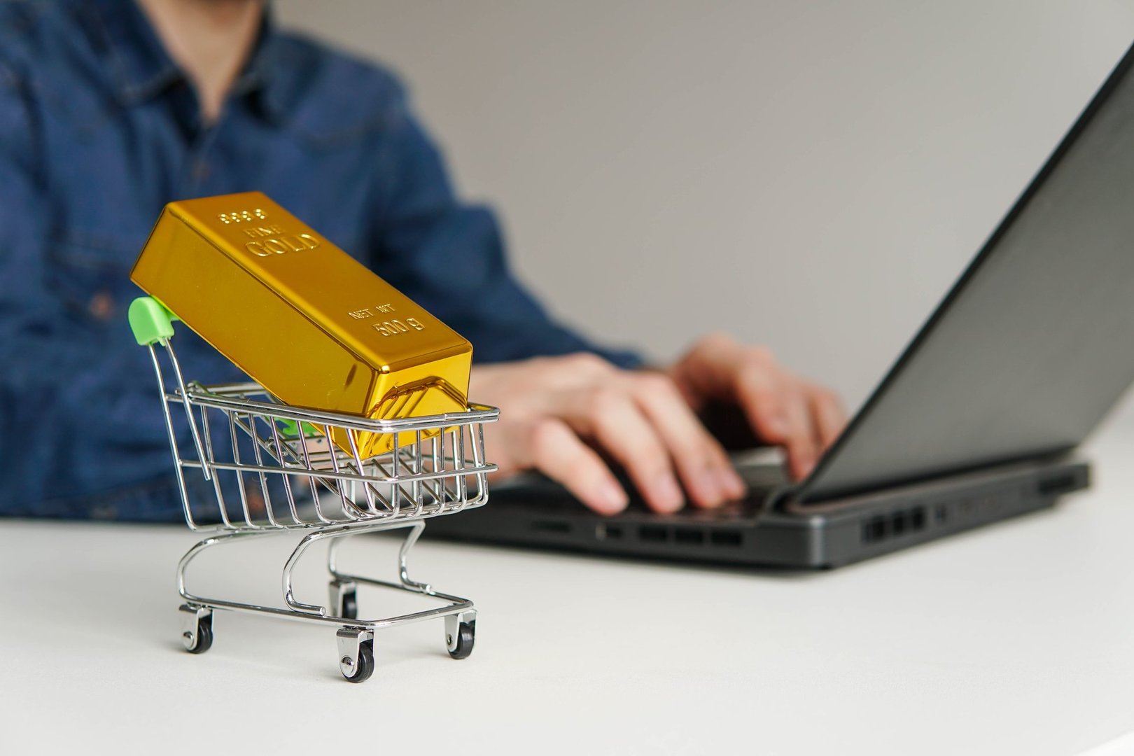 Small shopping cart with gold bar on the table. Man using laptop for online trading or buying gold bars for investment.