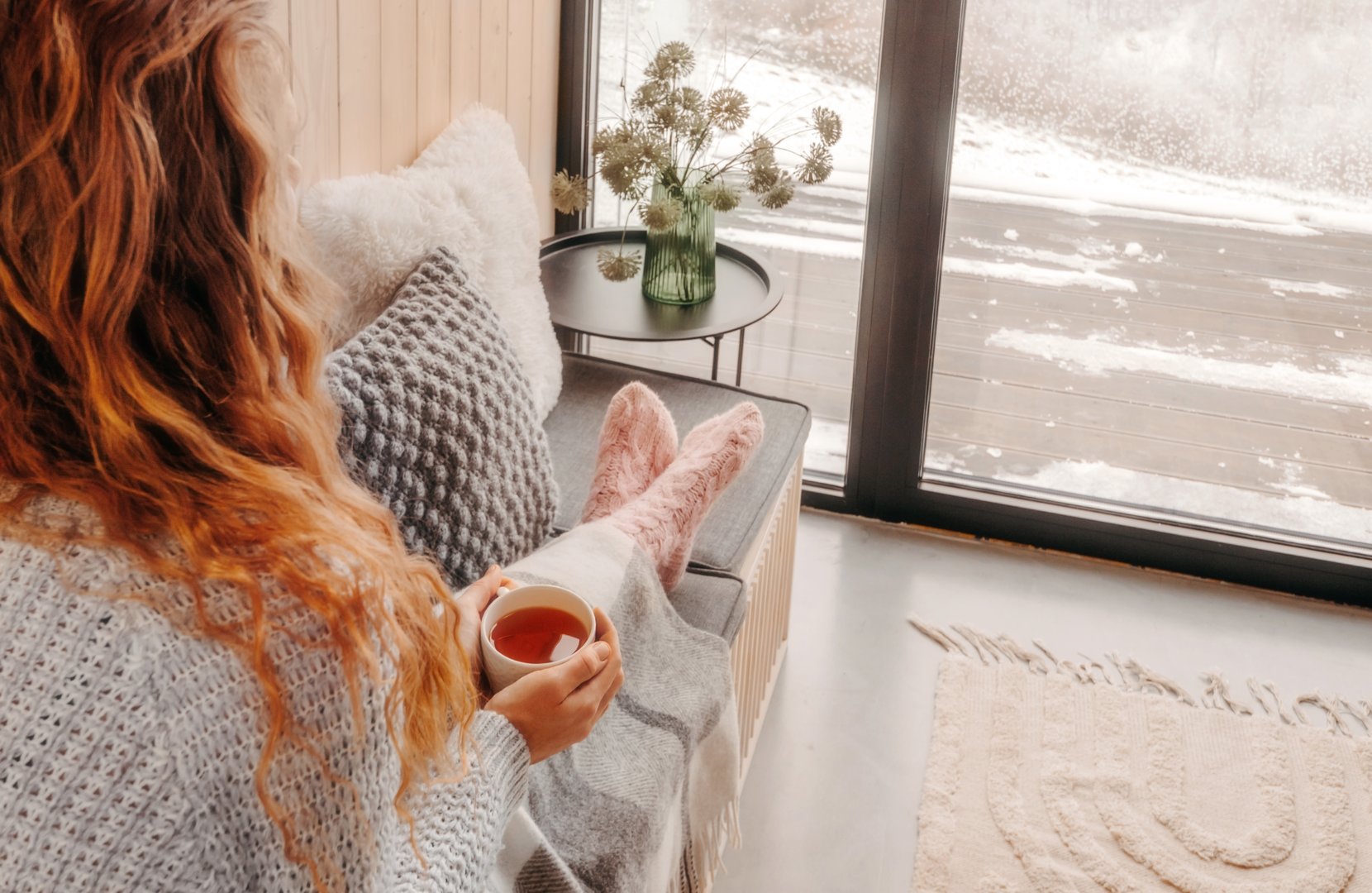 Woman sitting on sofa holding cup of tea, her legs are covered with cozy plaid blanket. Female enjoying slow life, looking through the window on winter day, spending weekend in rustic mountain cabin.