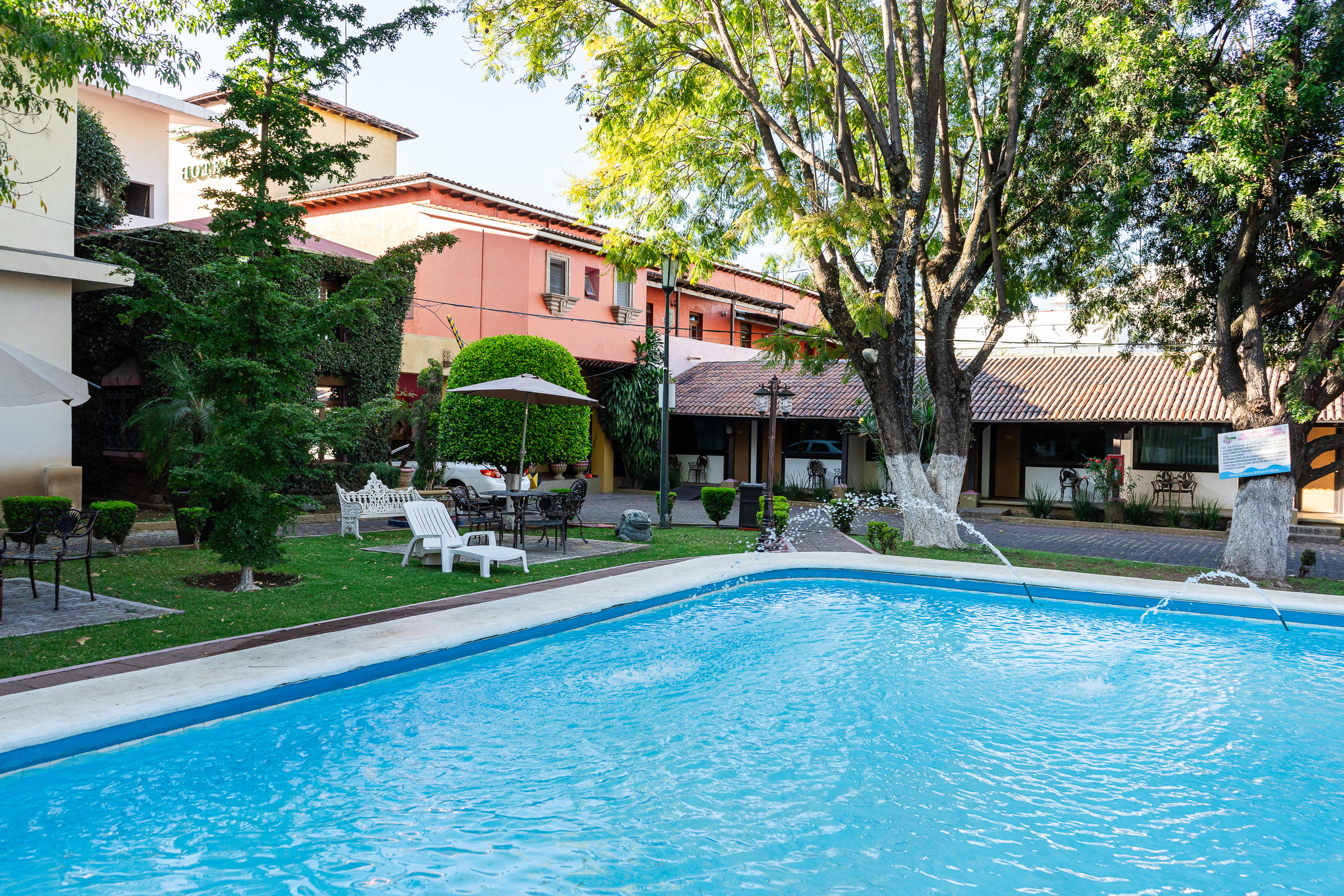 Outdoor pool with lounge chairs, umbrellas, and surrounding trees at a hotel, showing building exteriors nearby.
