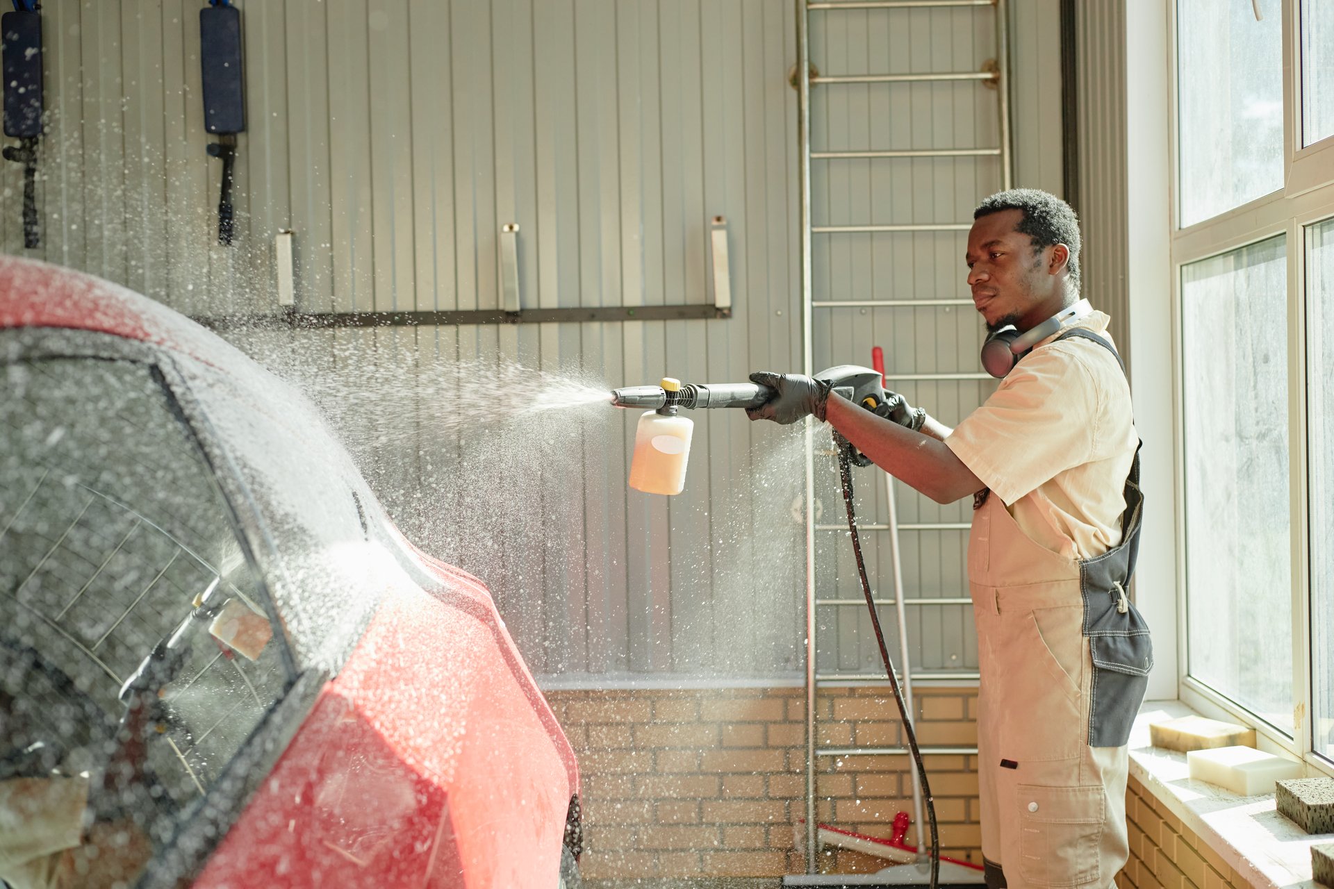 Black young adult man cleaning red car with high pressure washer in garage