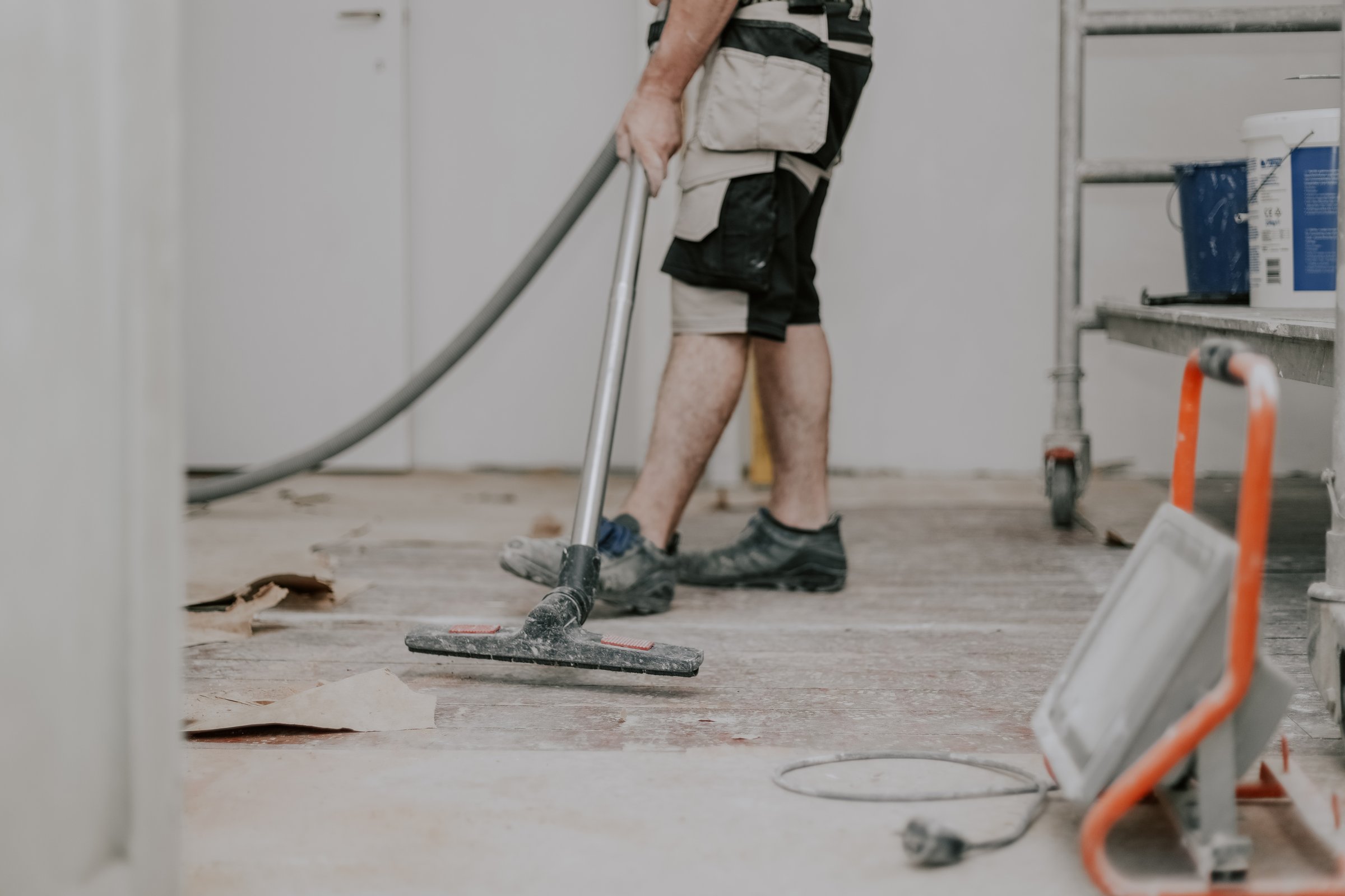 A young caucasian brunette man unrecognizable in work clothes on the left is vacuuming the floor with a construction vacuum cleaner in a room in an old house where renovations are underway, close-up view from the bottom with selective focus.Construction work concept.