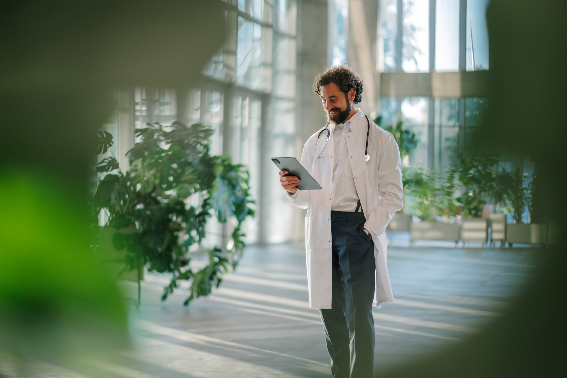 Smiling doctor walking through a modern hospital, using a digital tablet to connect and communicate in the healthcare environment