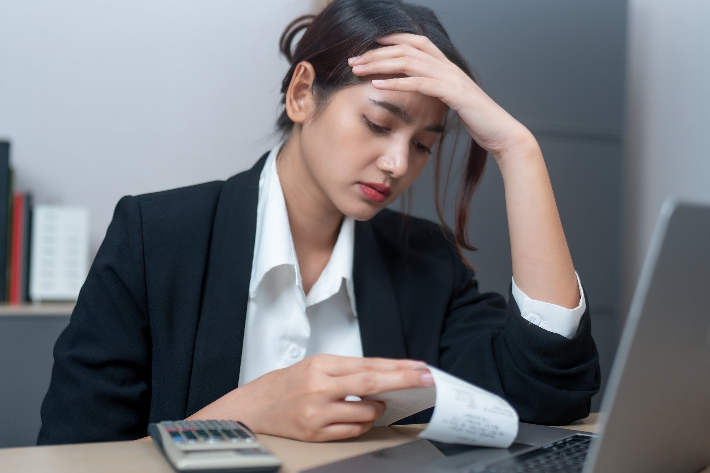 A stressed woman in black suit examines receipt while sitting at desk with laptop and calculator, reflecting financial concerns