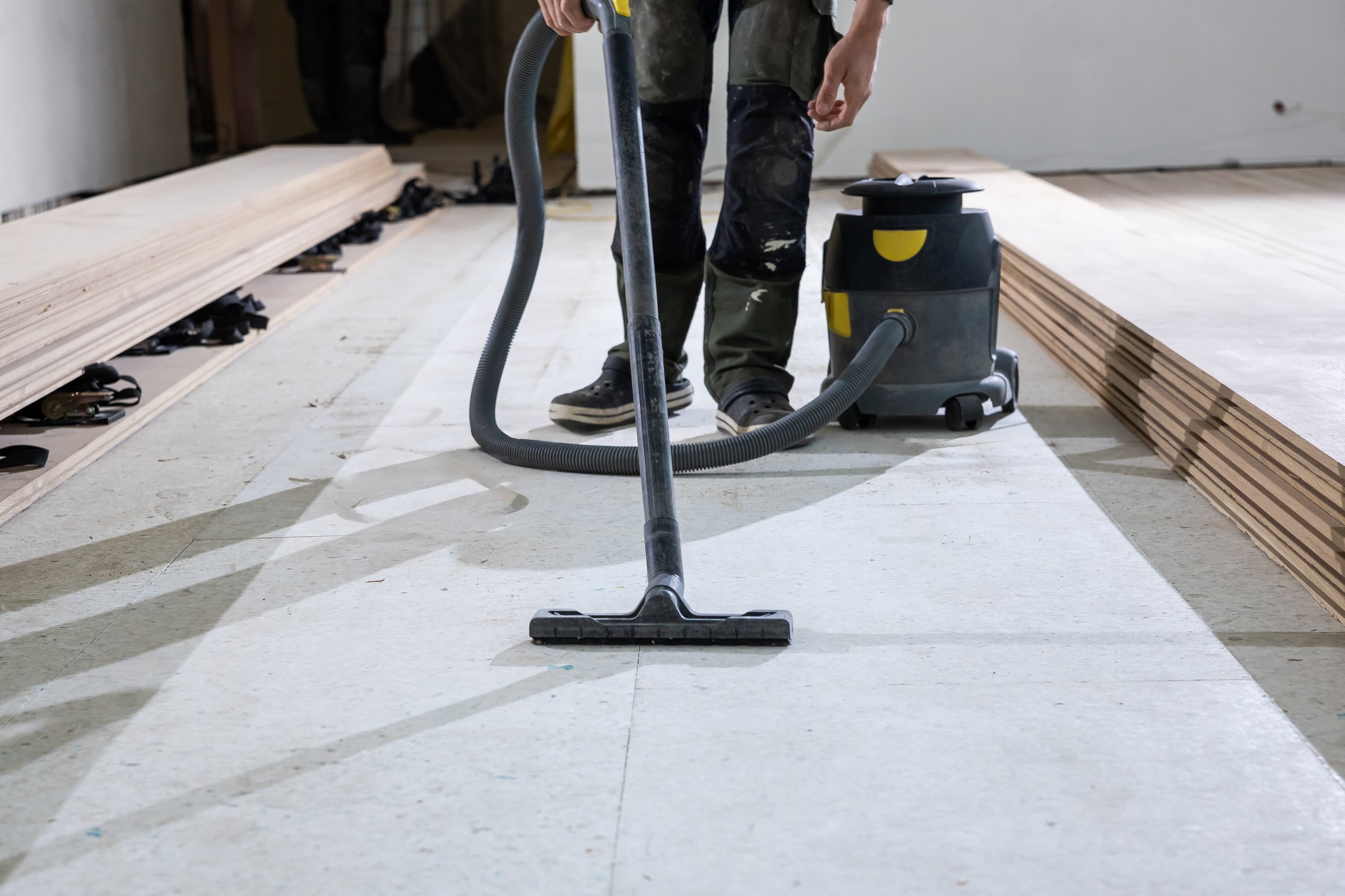 A worker uses a vacuum cleaner to clean a floor before installing wooden floorboards. Stacks of planks are visible, indicating a renovation project.