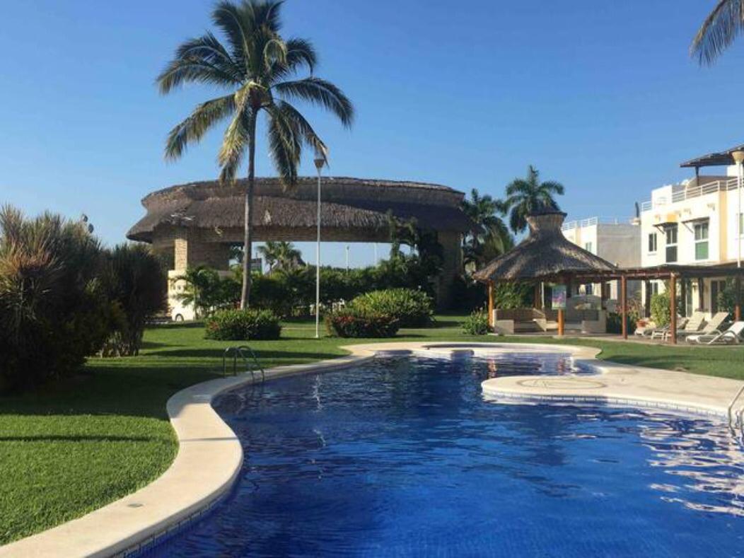 Tropical resort scene with a swimming pool, palm trees, and thatched-roof structures under a clear blue sky.