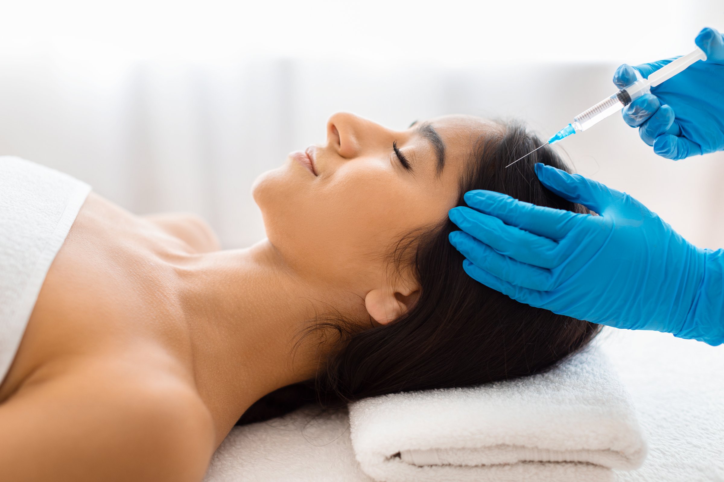 Hair loss, alopecia treatment. Unrecognizable beautician wearing medical gloves holding syringe, making scalp injections for smiling young indian lady, SPA beauty salon interior