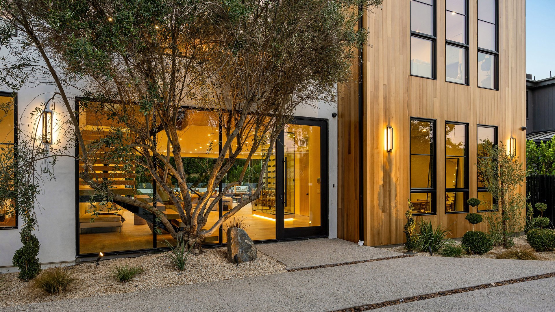 Modern house exterior with large windows and warm lighting, featuring a tree and landscaped garden in the foreground.
