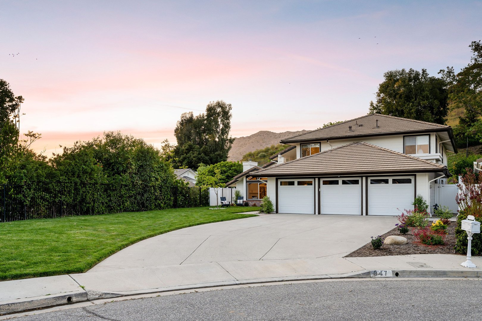 A suburban two-story house with a three-car garage, well-maintained lawn, and a scenic background at sunset.