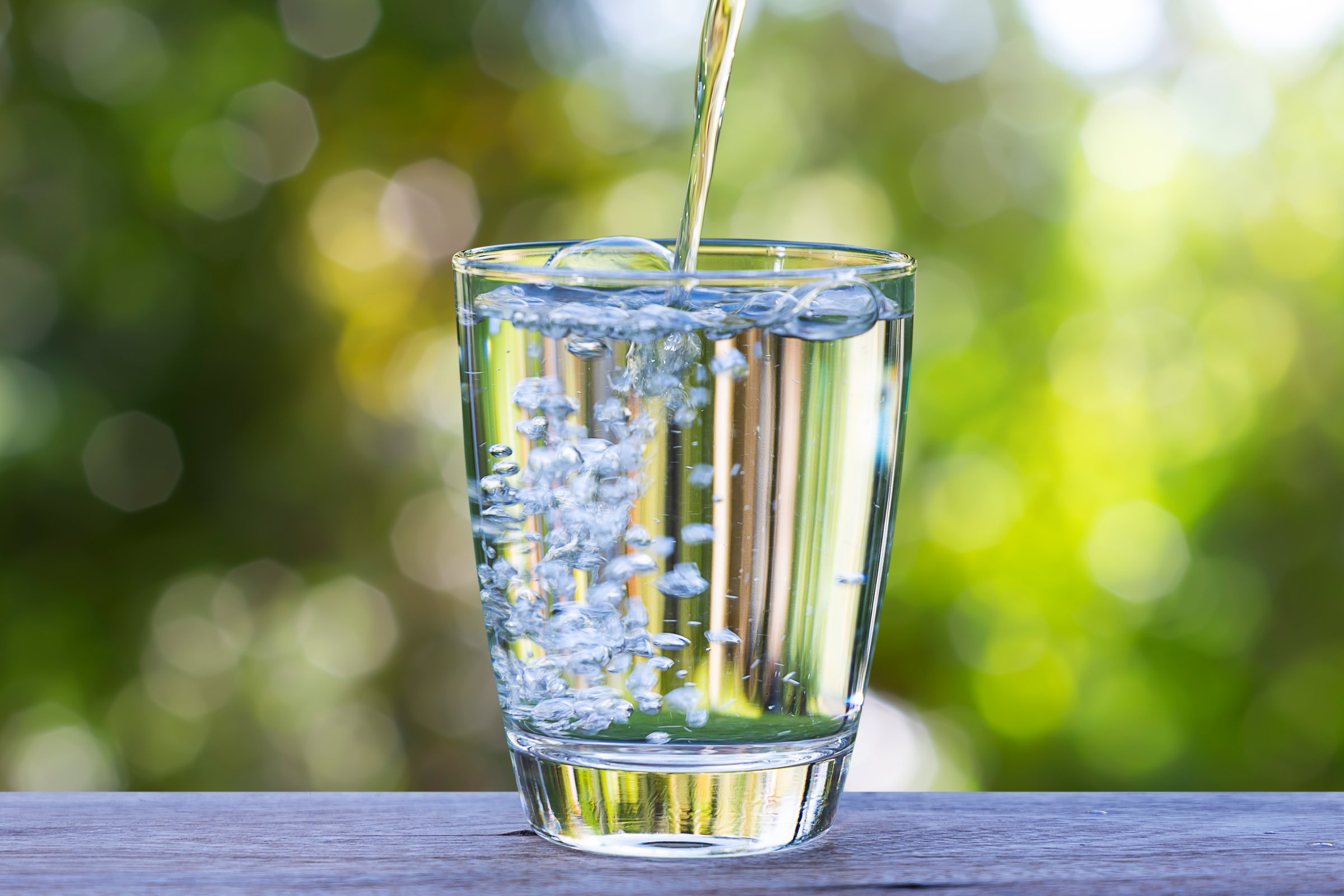 Pouring drinking water into glass on wooden table on blurred green nature bokeh background