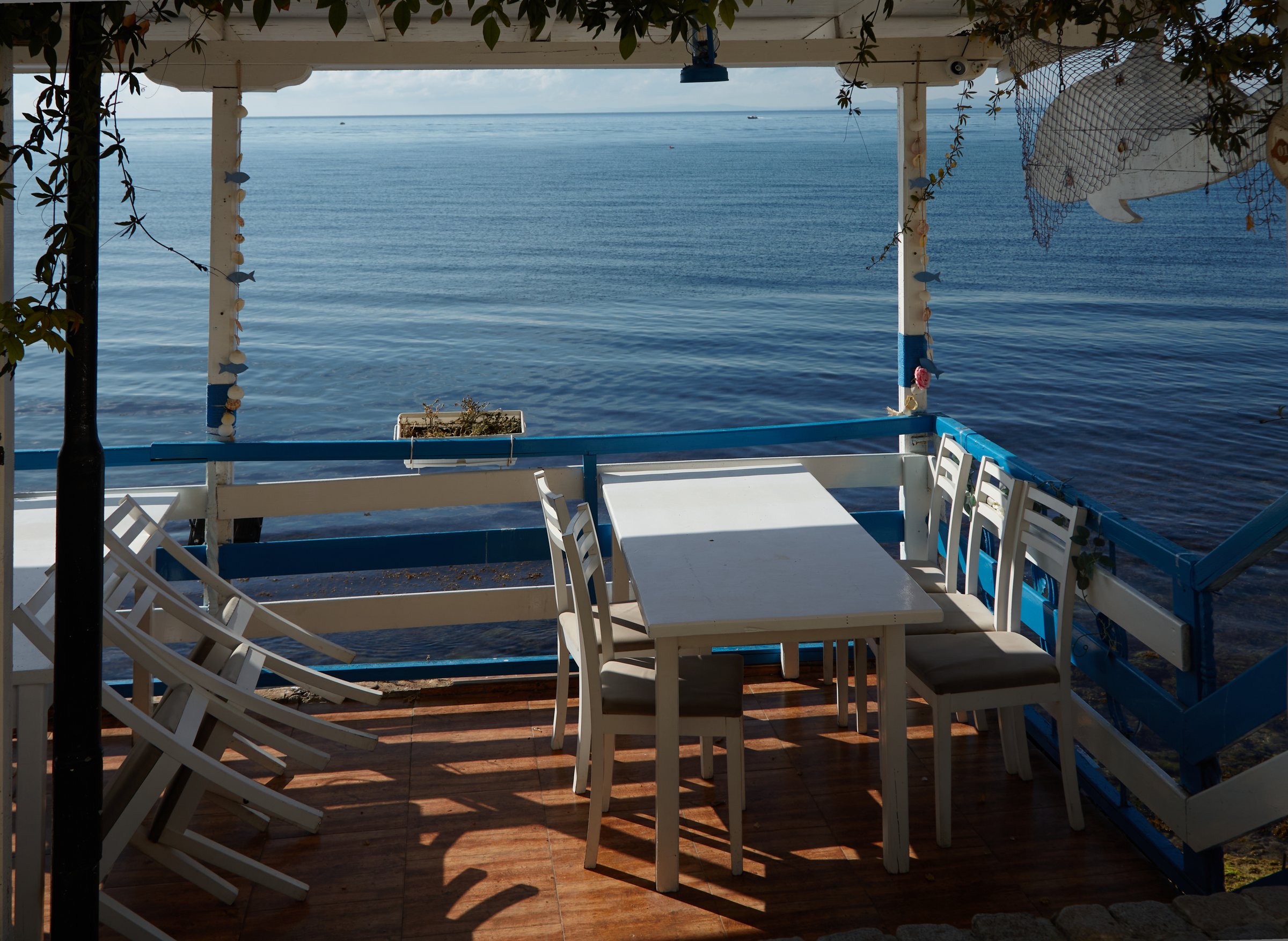 Waterfront dining space showcases white wooden tables and chairs, overlooking tranquil blue waters with distant boats, inviting guests to unwind and enjoy the peaceful ambiance of the seaside setting