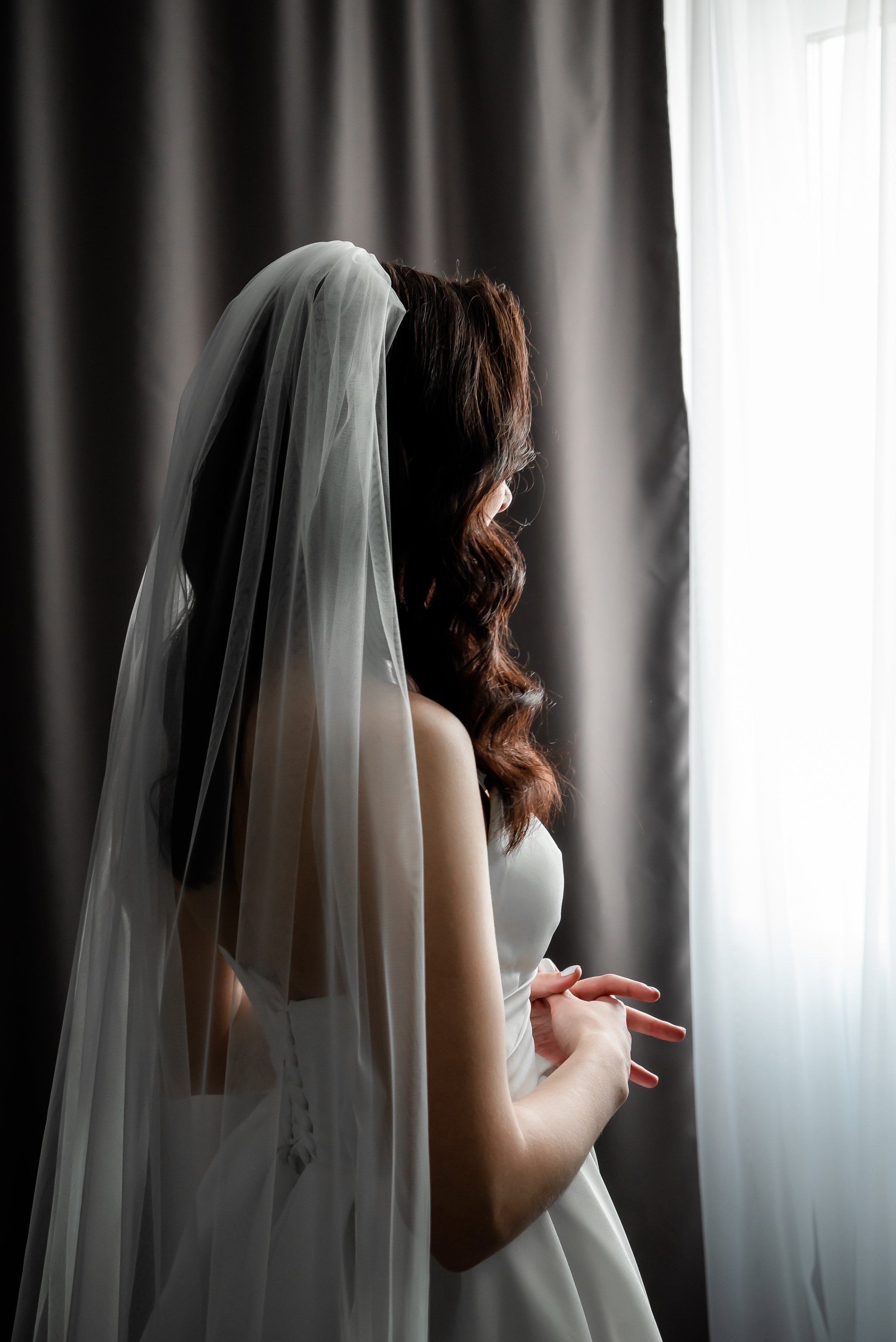 Bride stands in elegant wedding dress and veil, gazing out of the window, conveying anticipation and emotion before her special day. The serene moment captures her poised and reflective demeanor.