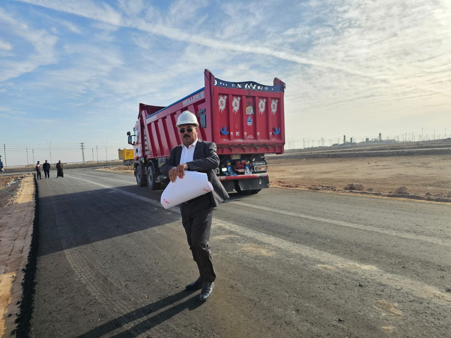 A man in a hard hat holds blueprints on a construction site with a red dump truck on a paved road.