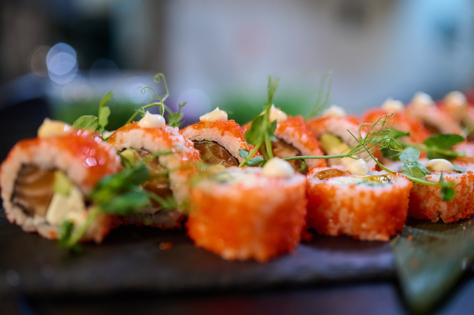 Close-up of delicious sushi rolls topped with tobiko, cream sauce, and microgreens, arranged on a black slate plate in a Japanese restaurant.