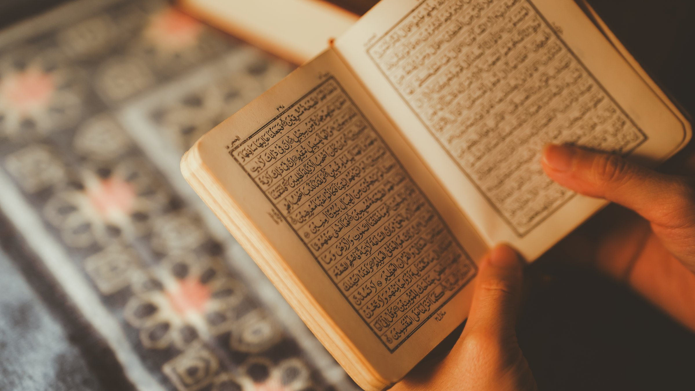 Close-up of a person's hands holding and reading the Quran, showing Islamic faith, spirituality, and religious devotion in soft lighting.
