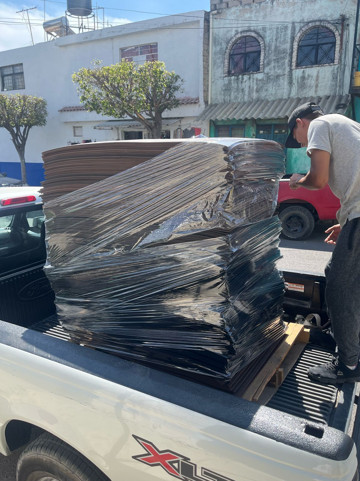 Stack of cardboard sheets wrapped in plastic on a truck bed next to a man in a cap adjusting the load on a sunny street.