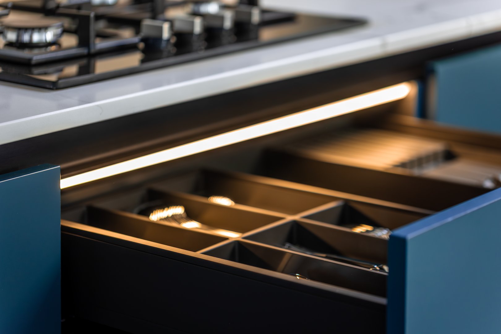 Pull-out drawers with organizers and lighting in a modern blue kitchen featuring a quartz countertop and gas hob.