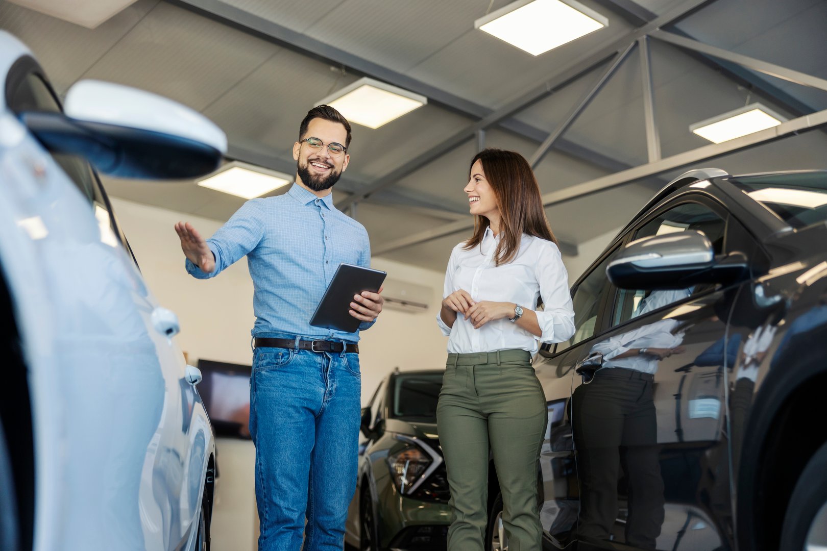Smiling friendly car salesman with tablet explaining car performances to a female customer at car salon.