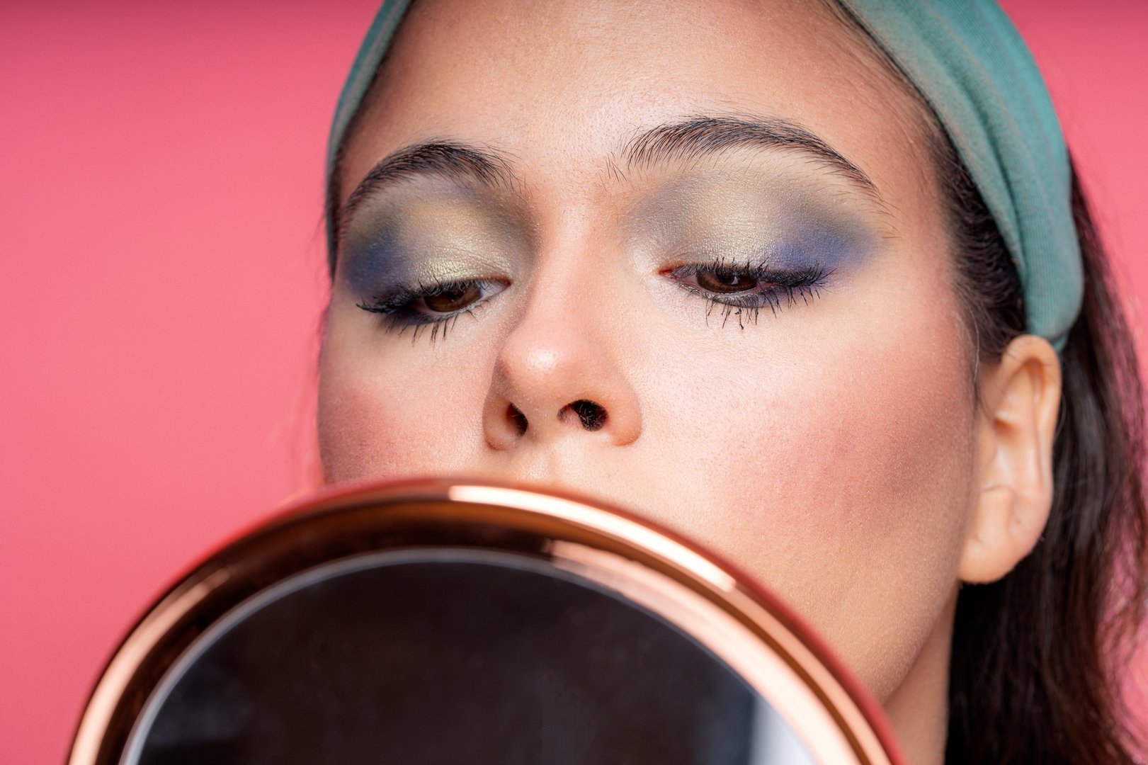 Close-up of a young woman with colorful eyeshadow looking at a mirror while applying makeup against a pink background