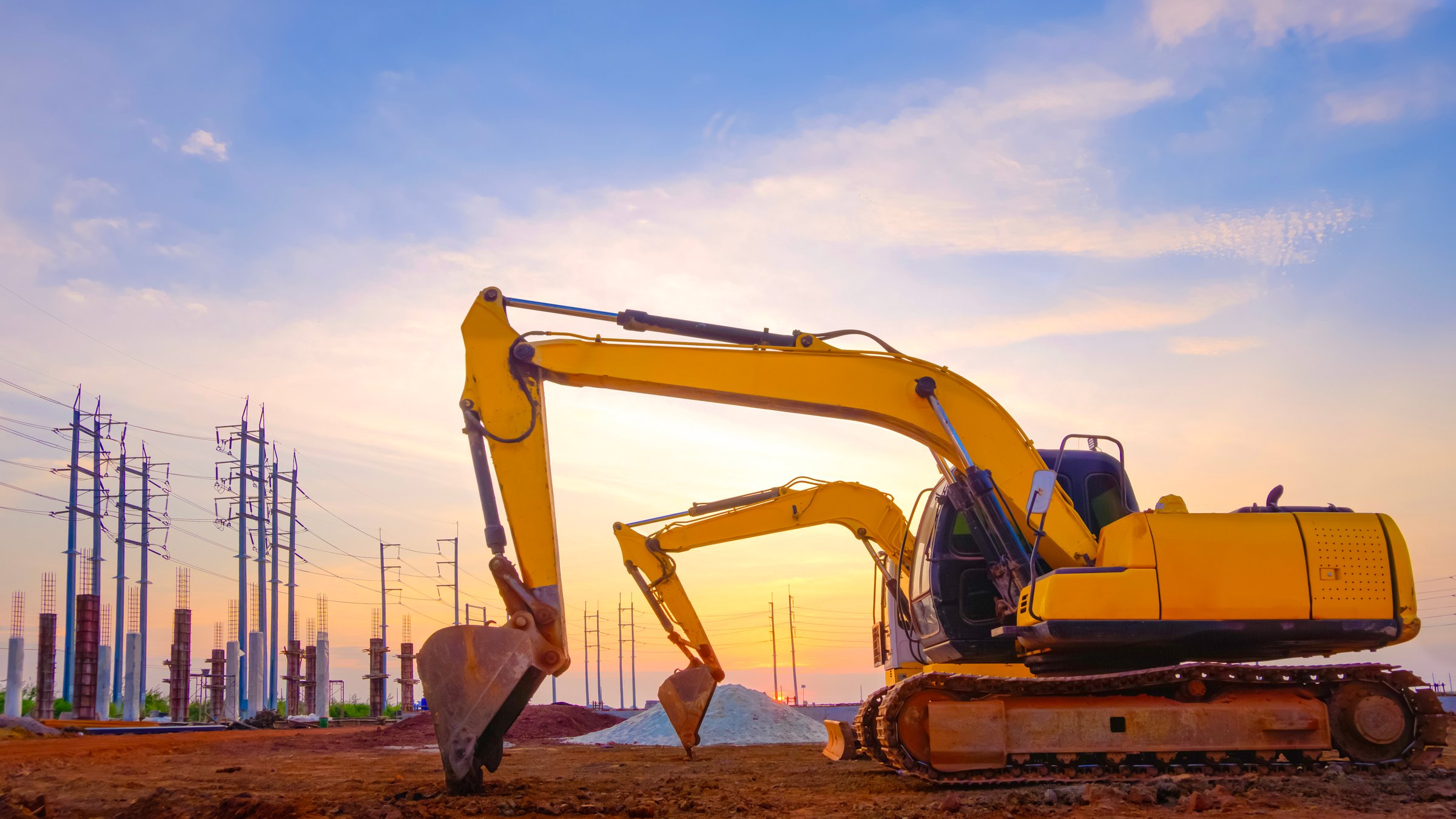 2 excavators parked on the ground in land filling area of new industrial factory construction site with row of electric poles against sunset sky background.