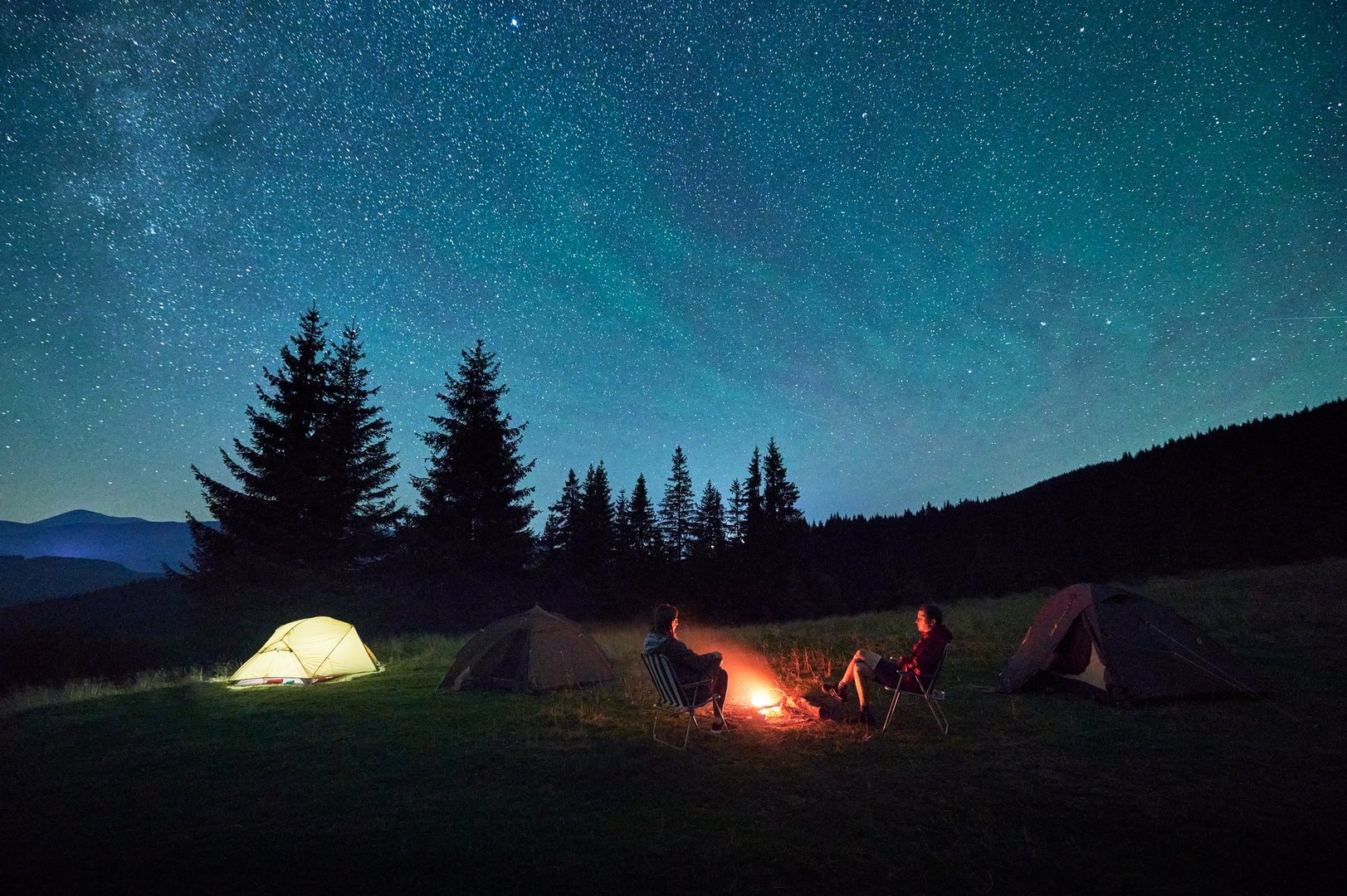 Silhouette of couple hikers resting by campfire under star-studded sky, near illuminated tents at campsite. Night camping in mountains, night sky create magical and peaceful atmosphere.