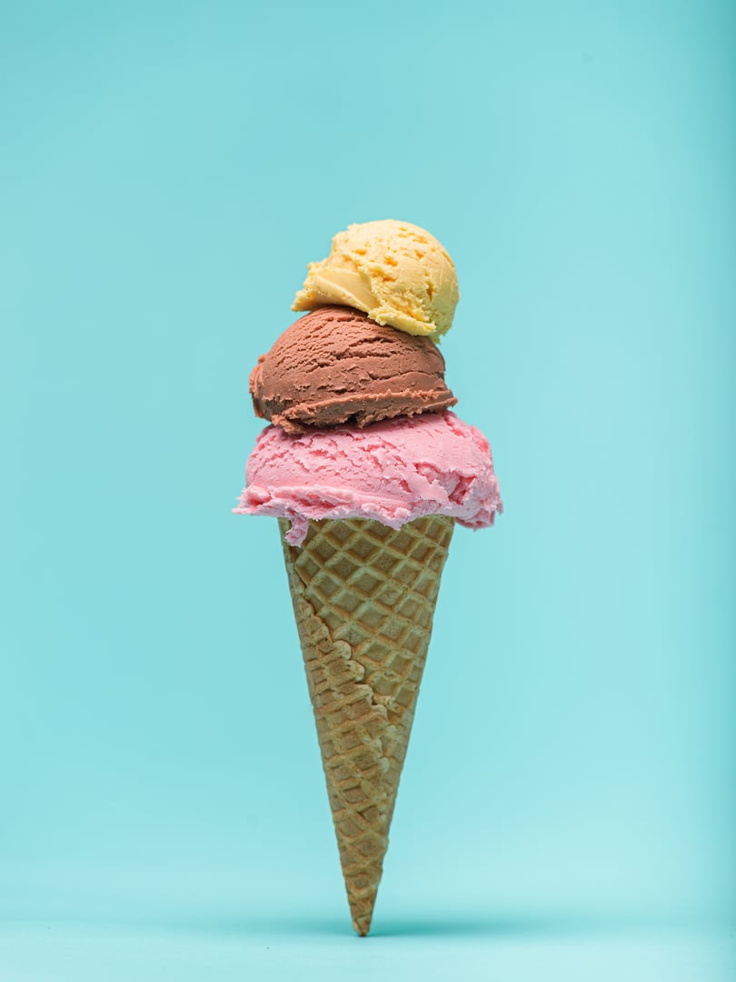 A waffle cone overflows with three scoops of ice cream: strawberry, chocolate, and vanilla. The vibrant colors pop against a pastel blue background